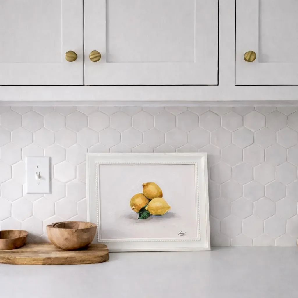 Kitchen with white hexagonal tile wall, white cabinets with gold knobs, a framed painting of three lemons, and wooden bowls on a cutting board.