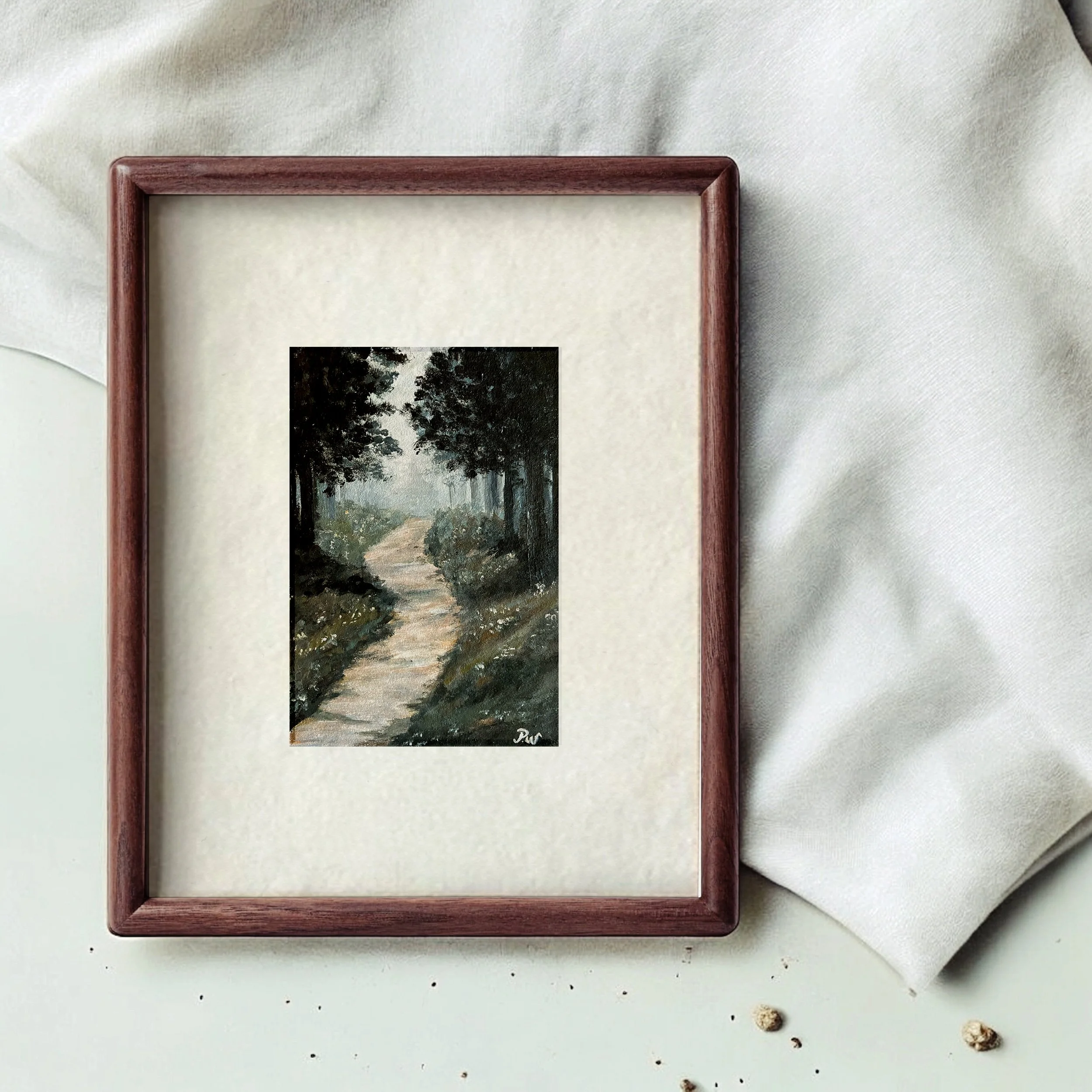 Miniature painting of a forest trail, framed and surrounded by white fabric, with some small objects on a white surface.