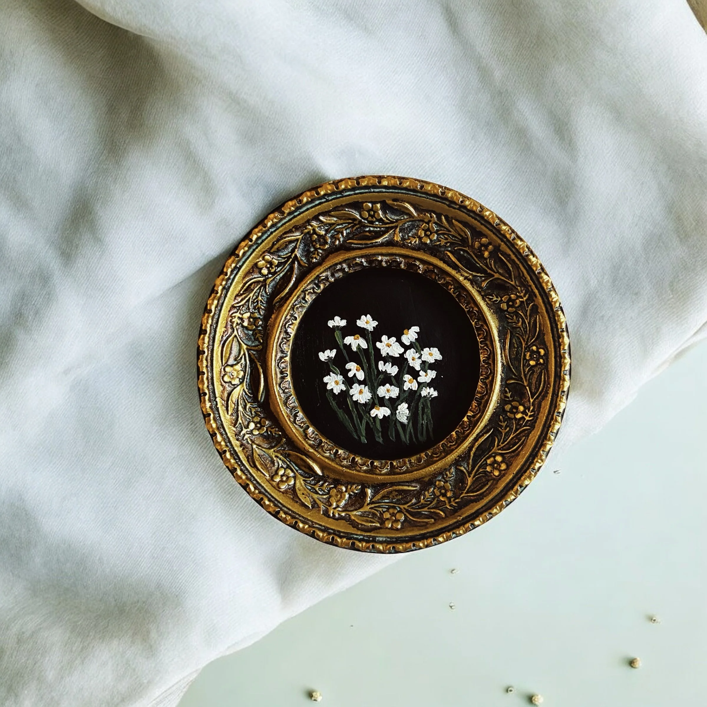Decorative golden oval frame with intricate floral patterns surrounding a black background with painted white daisies