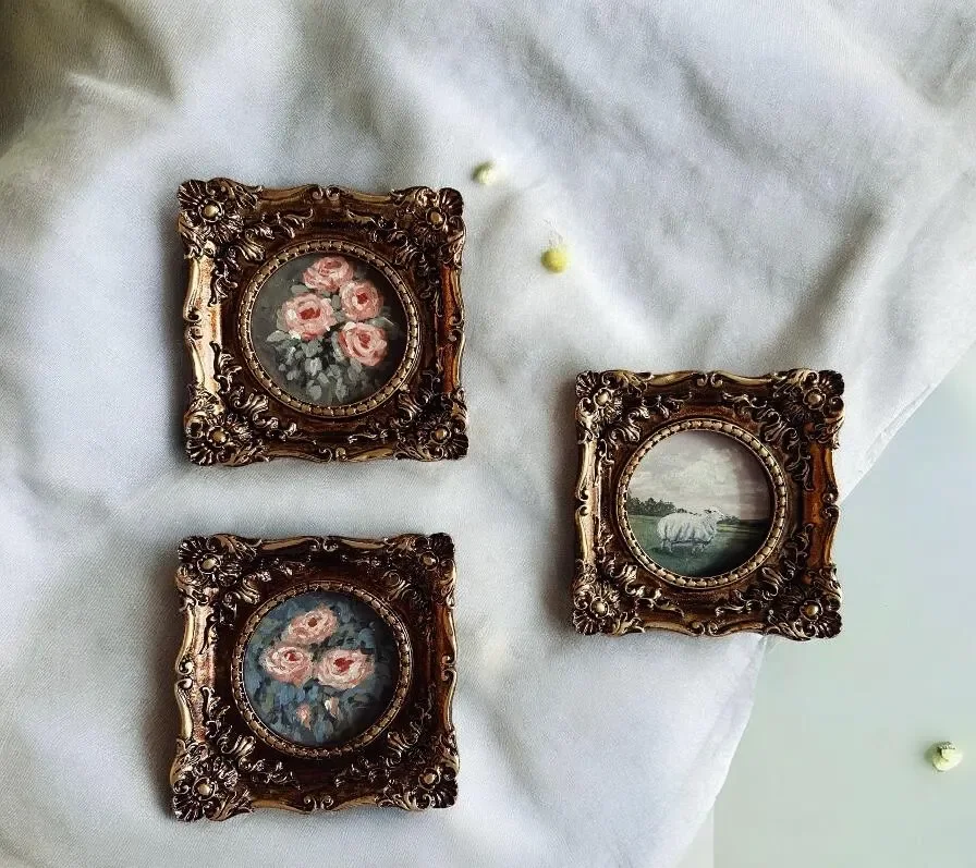 Three small vintage framed pictures on a white fabric surface. The top left and bottom left frames show floral paintings of pink roses, and the right frame shows a pastoral landscape with grazing sheep.