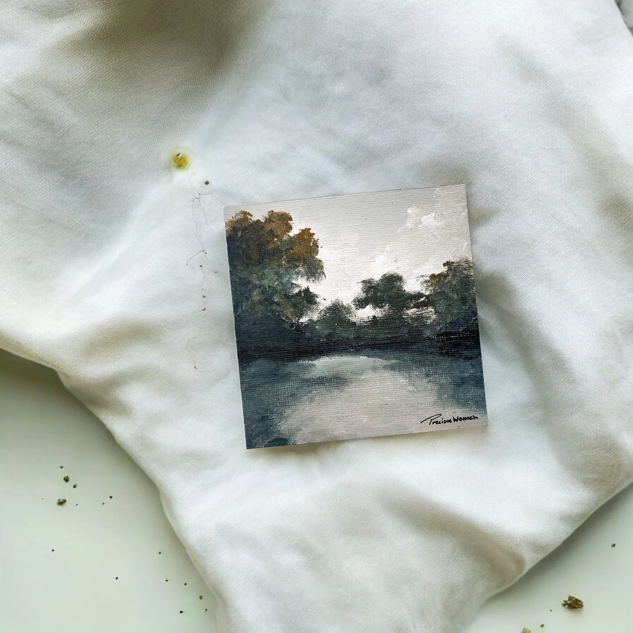 A small landscape painting of trees over water, signed 'Precious Wettach', placed on white fabric.