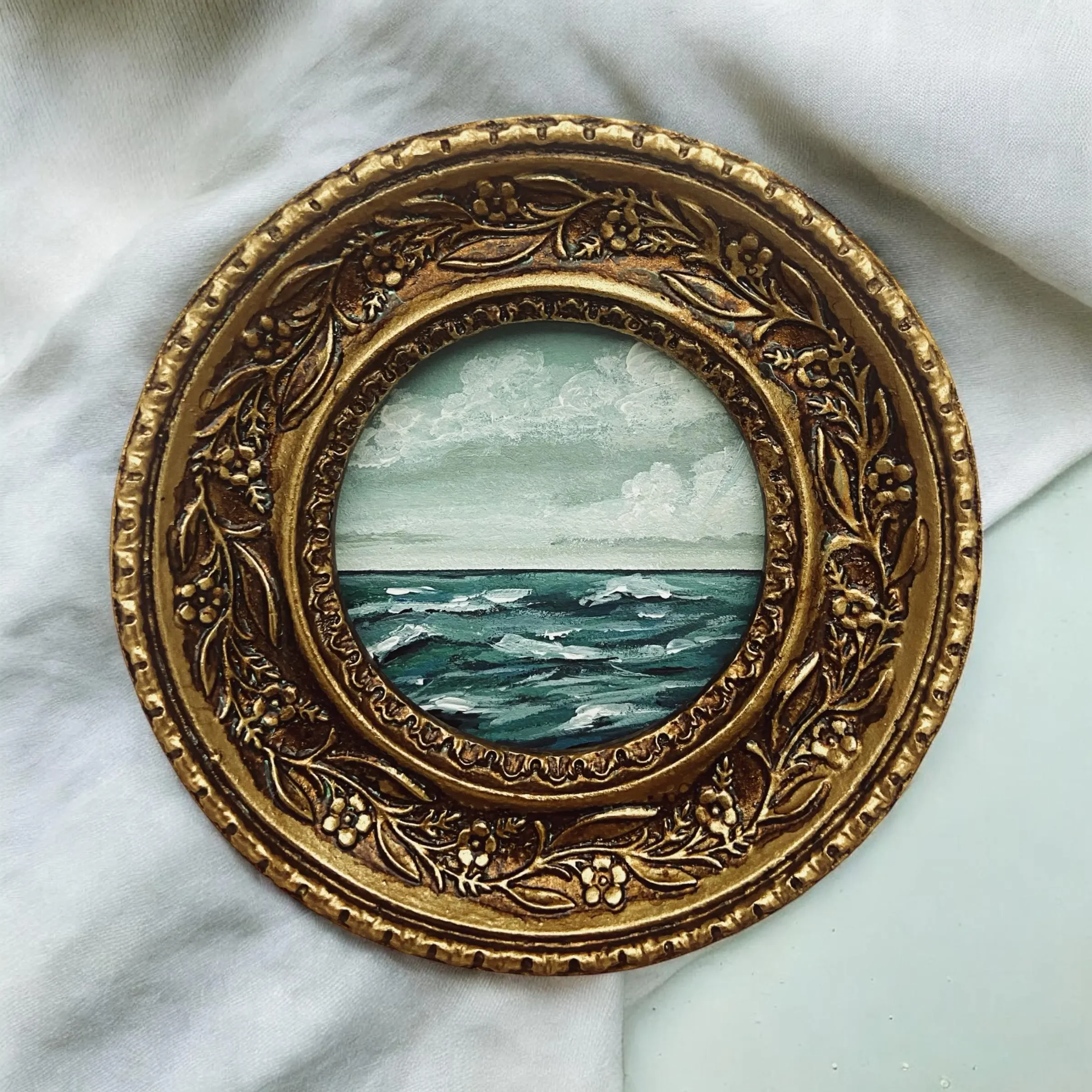 Decorative oval picture frame with floral and leaf designs, containing a painting of an ocean scene with waves, clouds, and sky.
