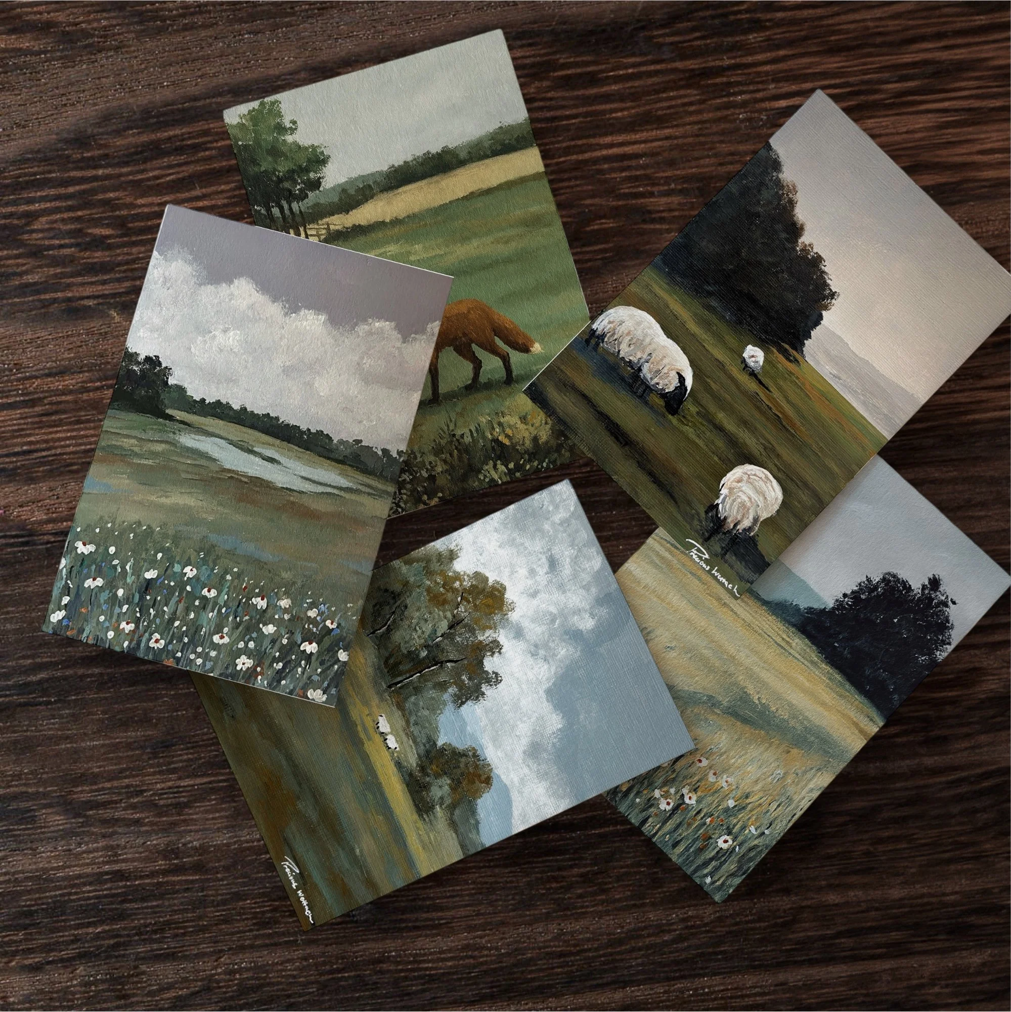 Collection of five landscape oil paintings depicting countryside scenes with fields, trees, and grazing sheep, placed on a wooden surface.