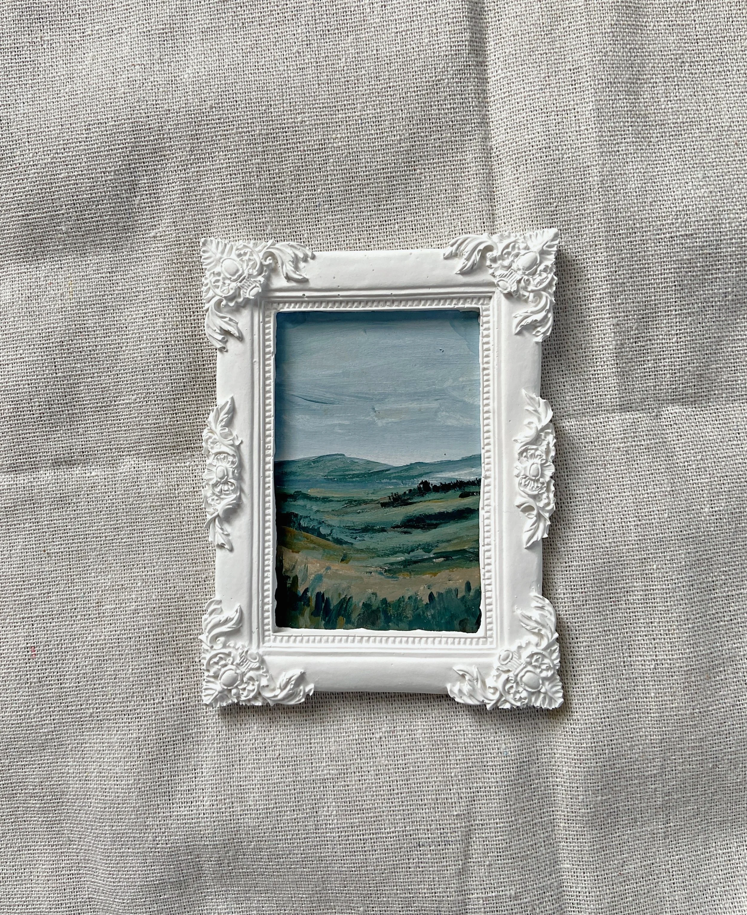 Small landscape painting of rolling hills and sky, framed in an ornate white frame, displayed on a beige fabric background.