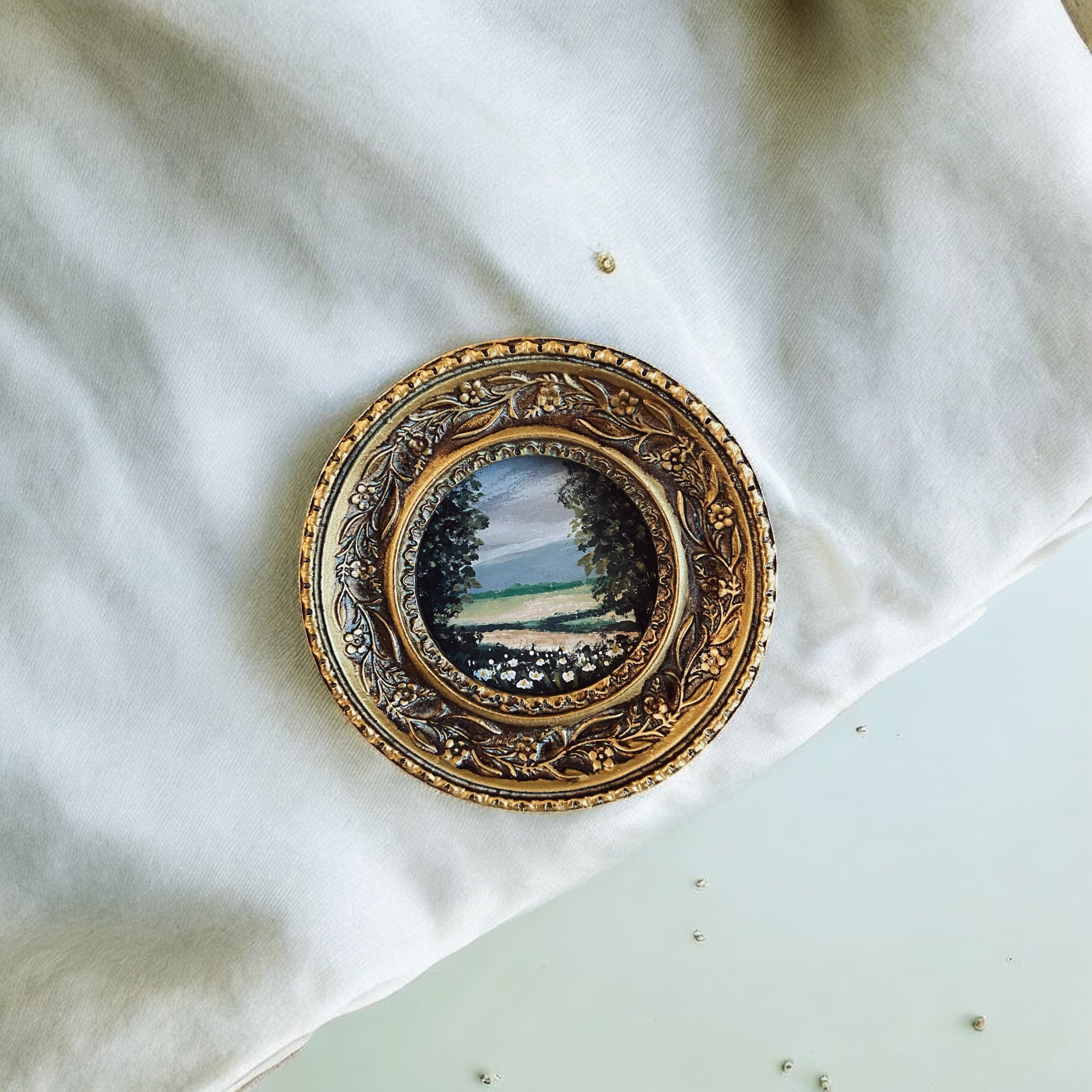 A decorative brooch with a landscape miniature design, resting on a white fabric surface.