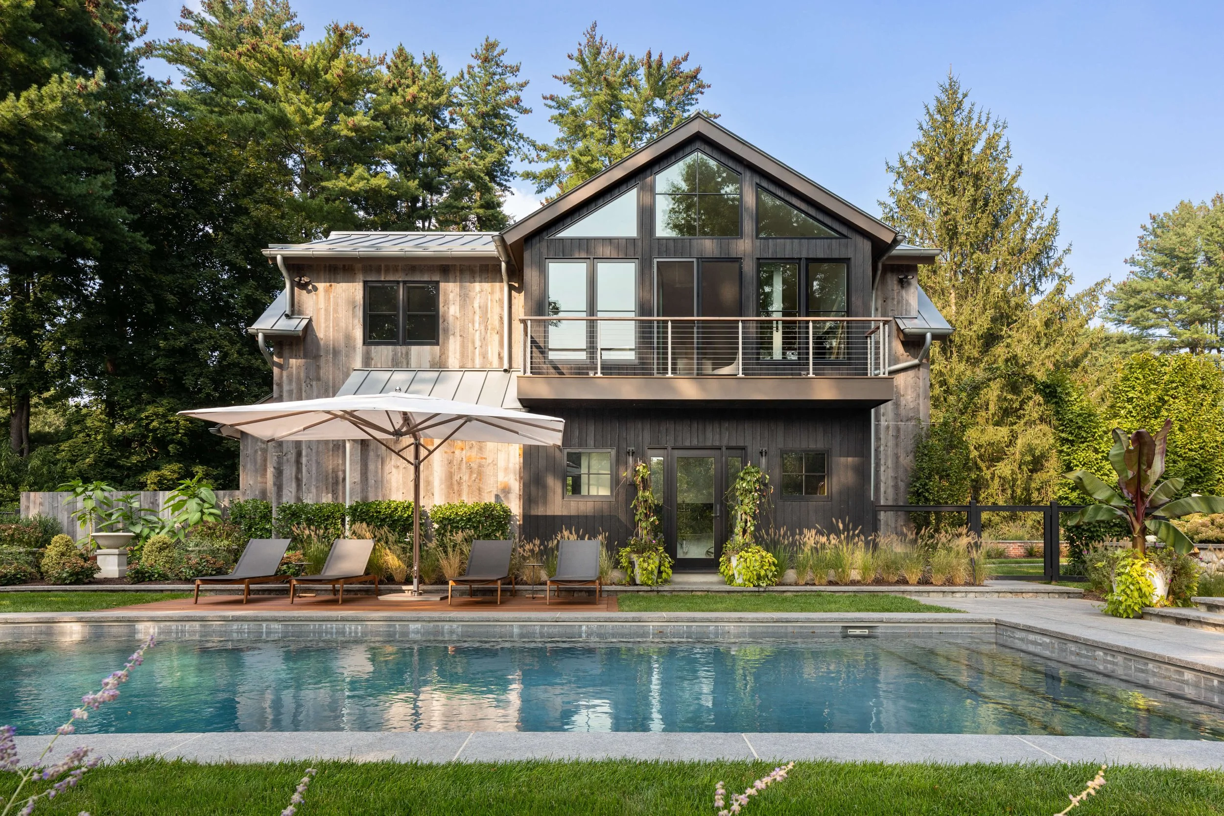 A modern two-story house with dark wood siding, large windows, and a balcony. In front, there is a swimming pool surrounded by lounge chairs, an umbrella, and landscaped greenery.