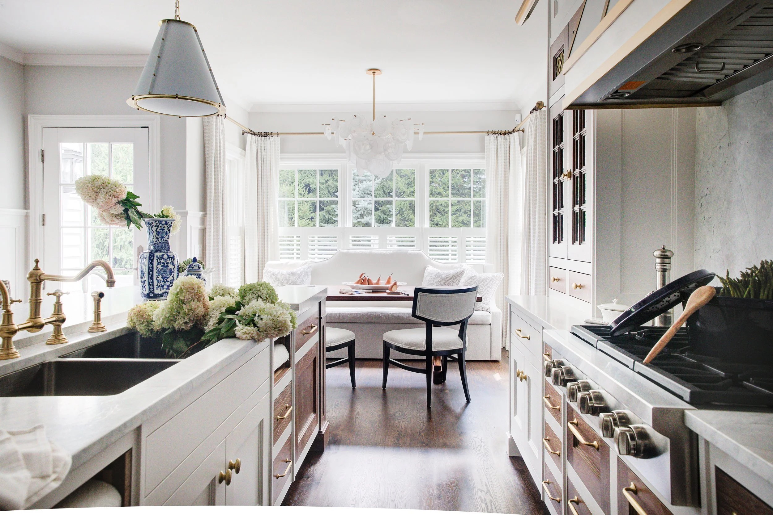 Bright kitchen with white cabinets, dark wood floors, and large windows; floral arrangements on the countertop, a stove with utensils, and a sitting area with chairs and a sofa in the background.