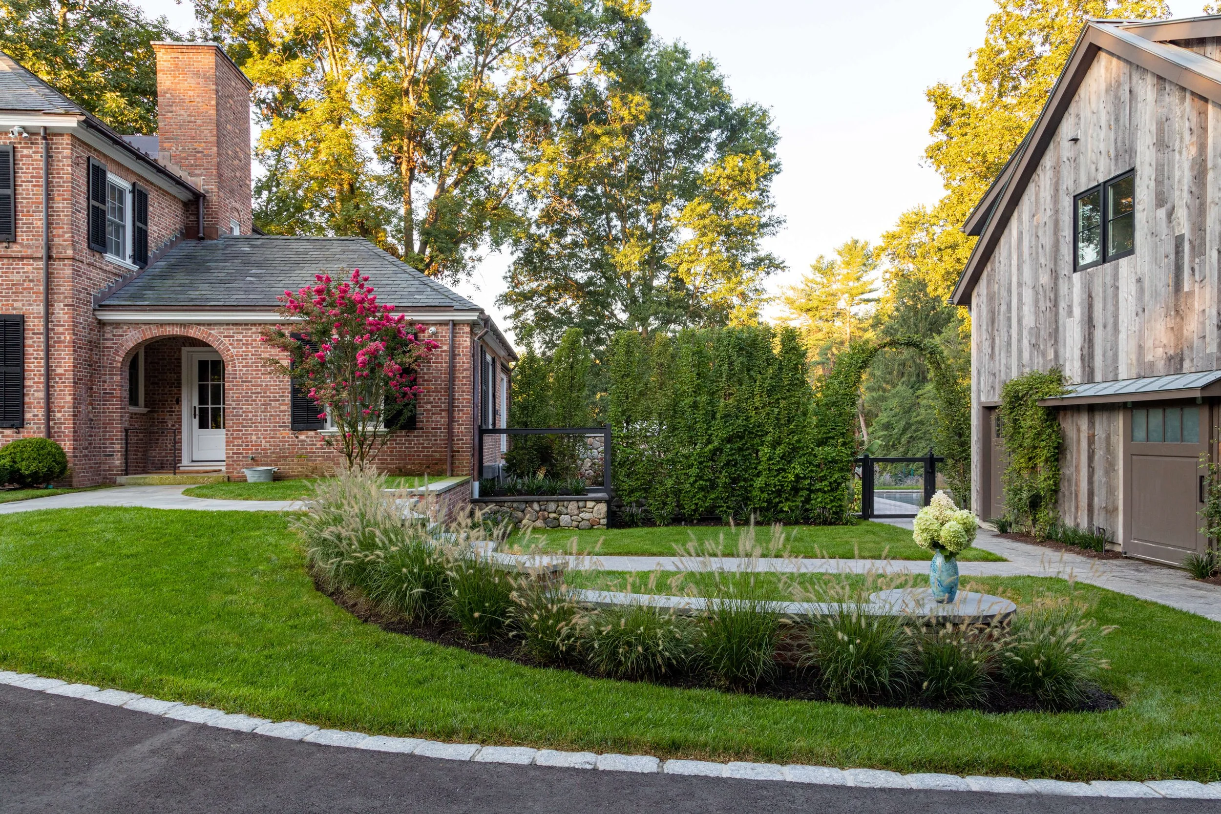 A well-maintained front yard with a curved driveway, green grass, a flowering pink tree, ornamental grasses, and a vase with white flowers on a concrete pad, bordered by bushes and trees, with a brick house on the left and a wooden garage on the right.