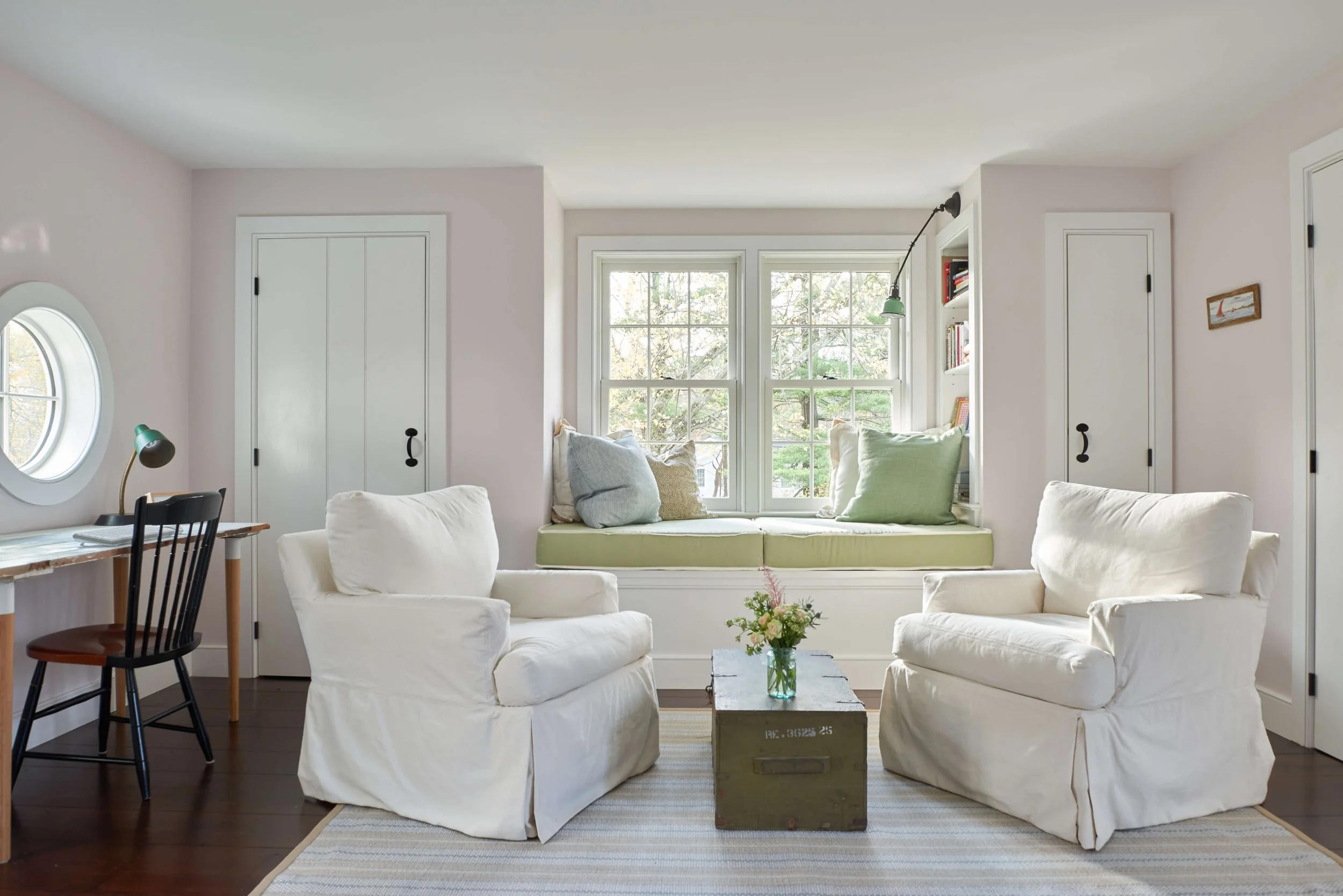 Living room with two white armchairs, a window seat with green cushions, large window, ladder shelf with books, small coffee table with flowers, dark wood flooring, side desk with black chair and green desk lamp, circular window, and pastel walls.