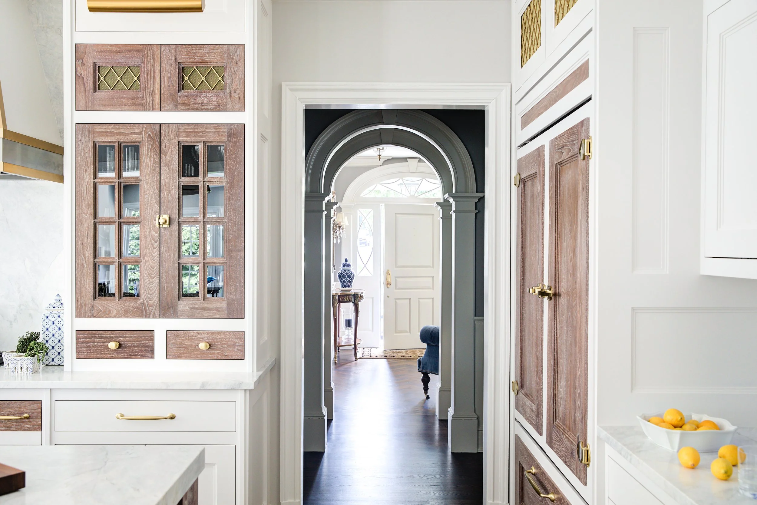 View through a kitchen doorway into an entryway with a front door, a small table with a blue vase, and a window with sunlight.