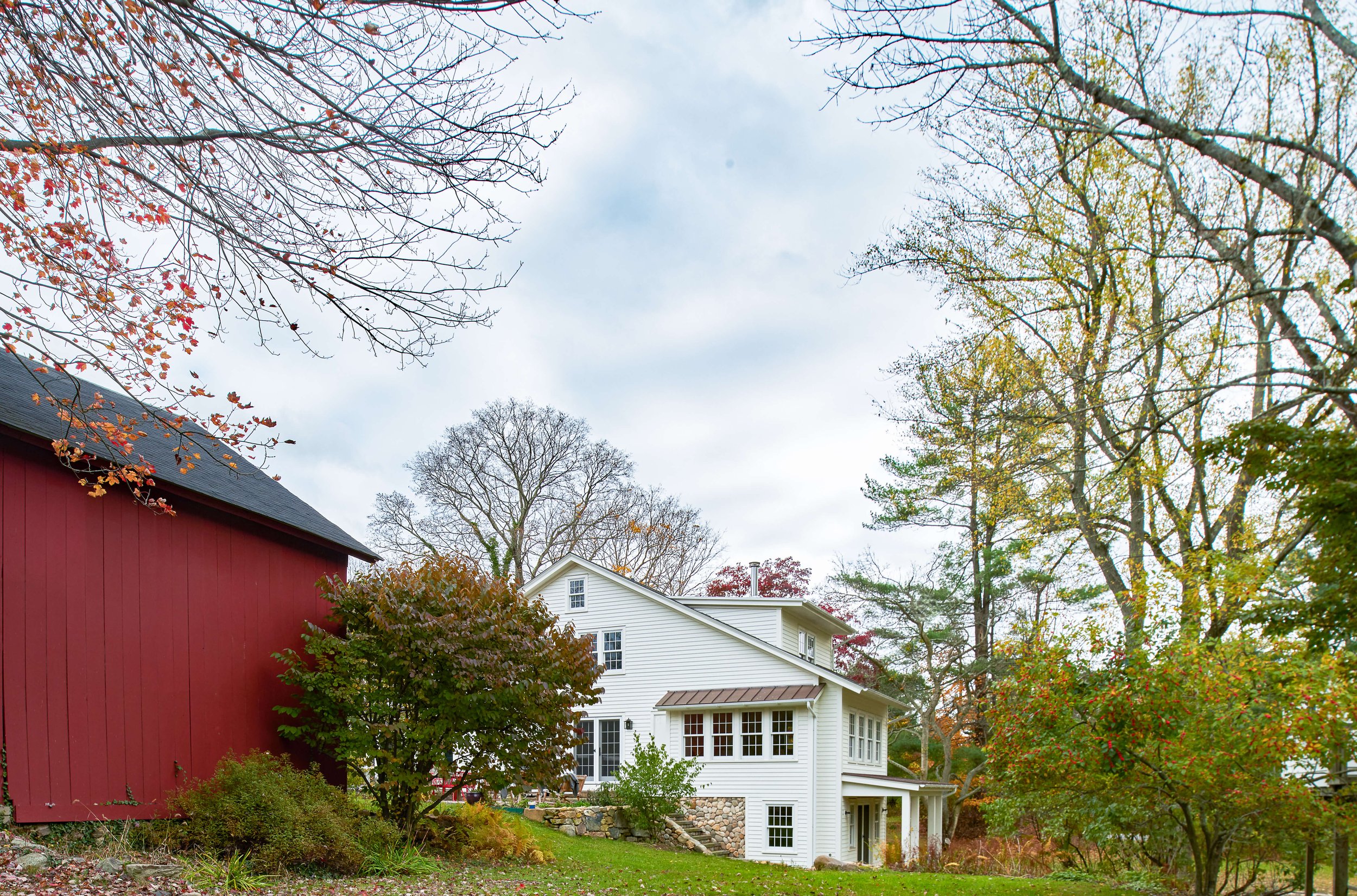 View of a white house with a porch in a yard surrounded by trees with autumn foliage, and a red barn on the left, under an overcast sky.
