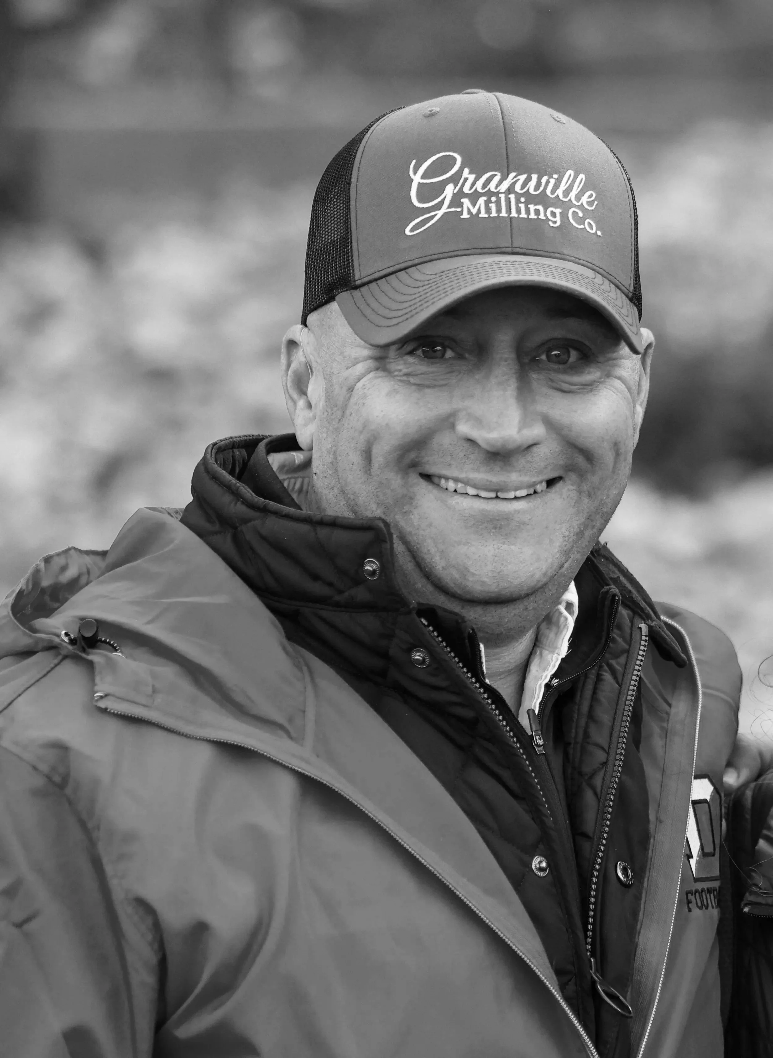 A smiling man wearing a cap with the words "Granville Milling Co." and a jacket. The background is blurred.