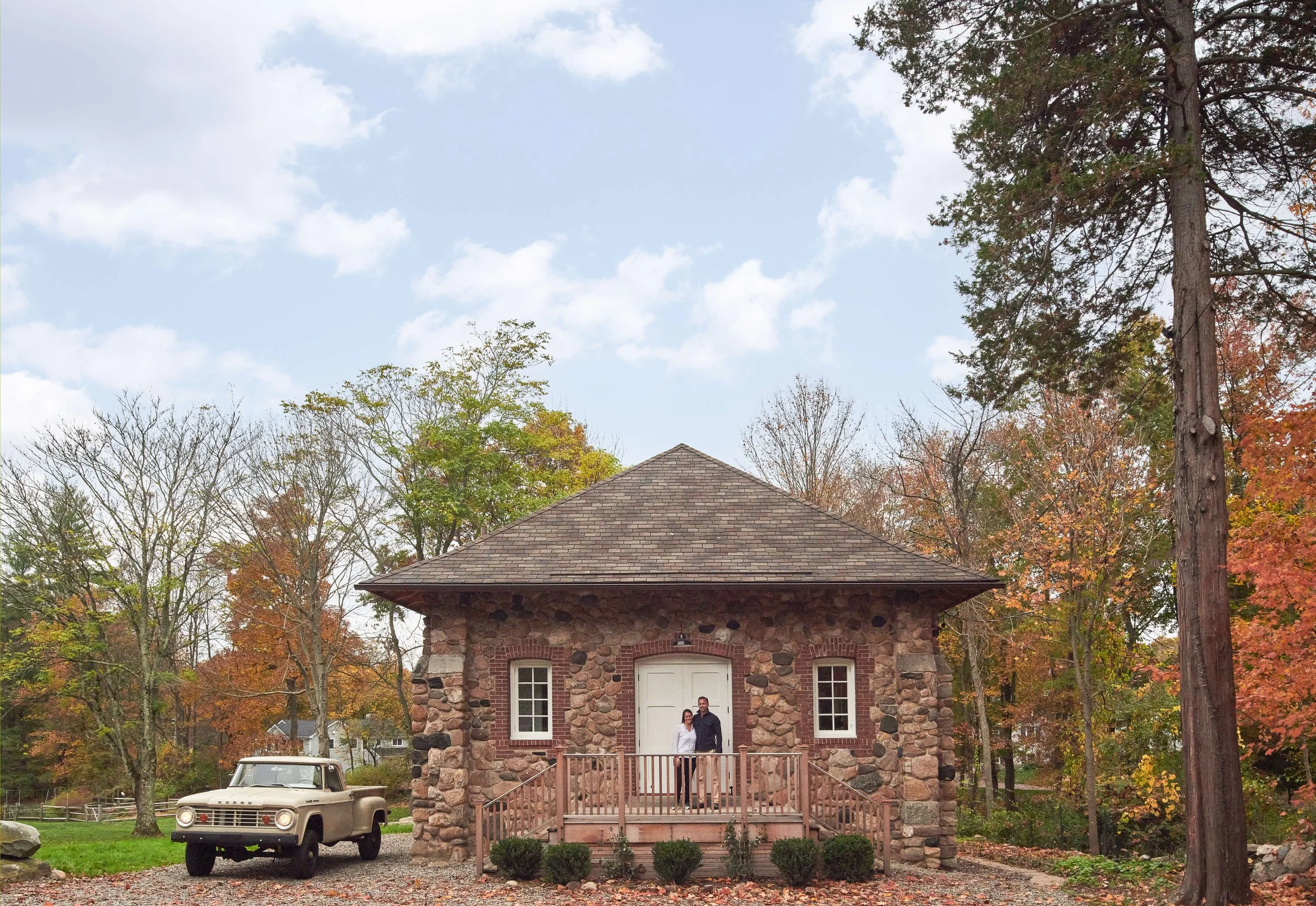 A small stone building with a porch, two people standing on the porch, a vintage pickup truck parked nearby, surrounded by trees with autumn foliage.