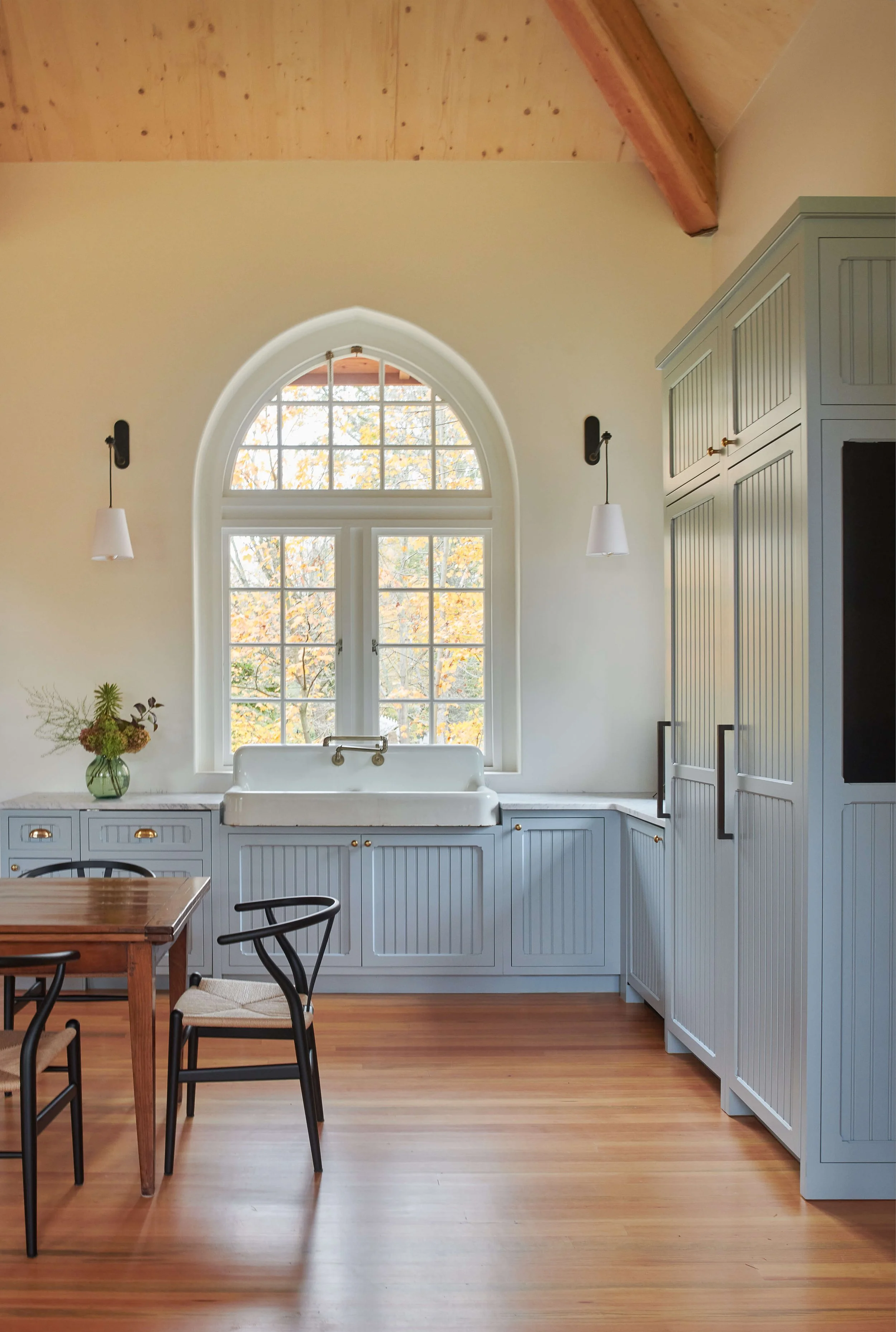 A bright kitchen with a large arched window, light blue cabinetry with gold handles, a farmhouse sink, a wooden dining table with black chairs, a green vase with flowers, and light wood flooring.