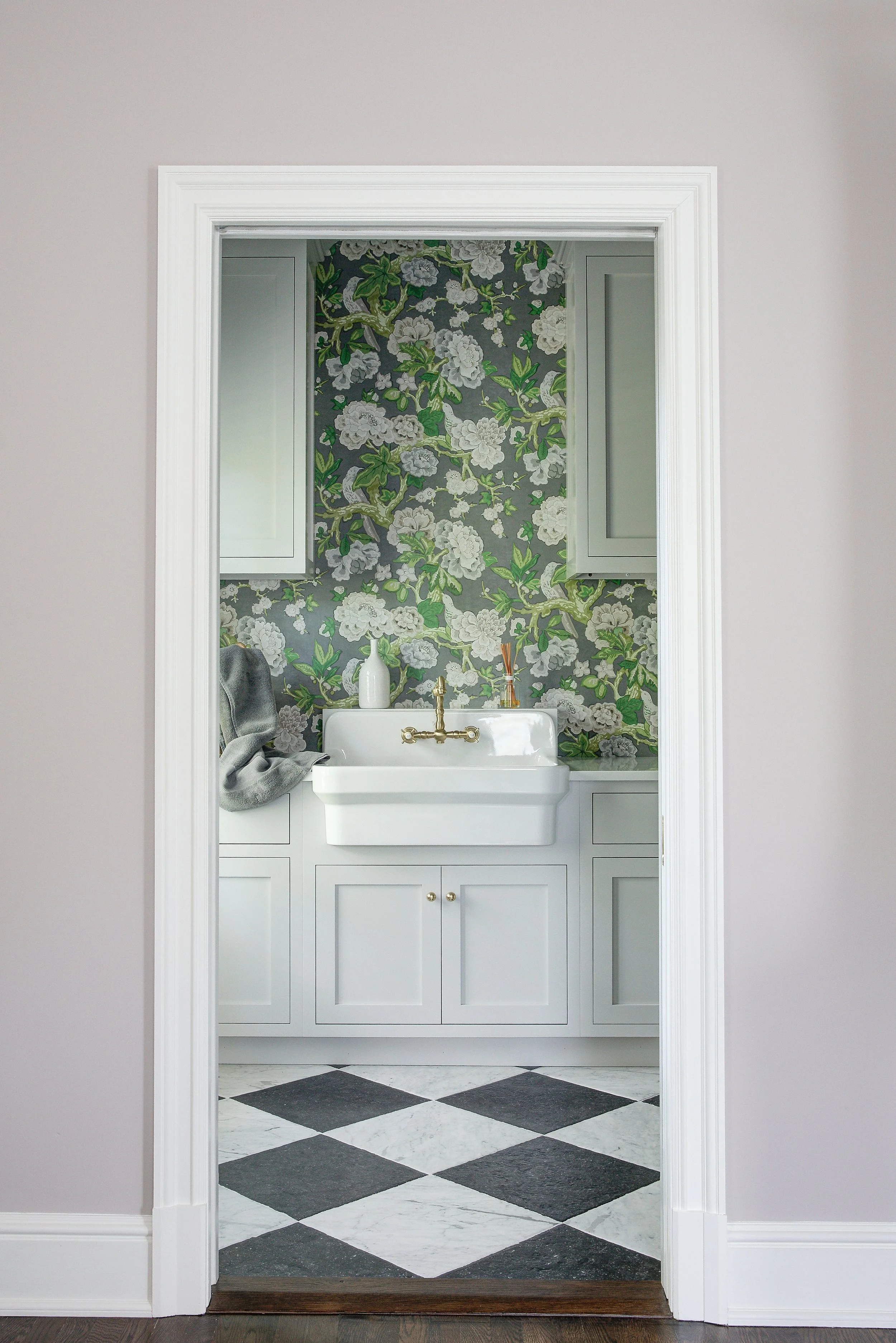View of a laundry or utility room with floral wallpaper, white cabinetry, and a white sink with brass fixtures.