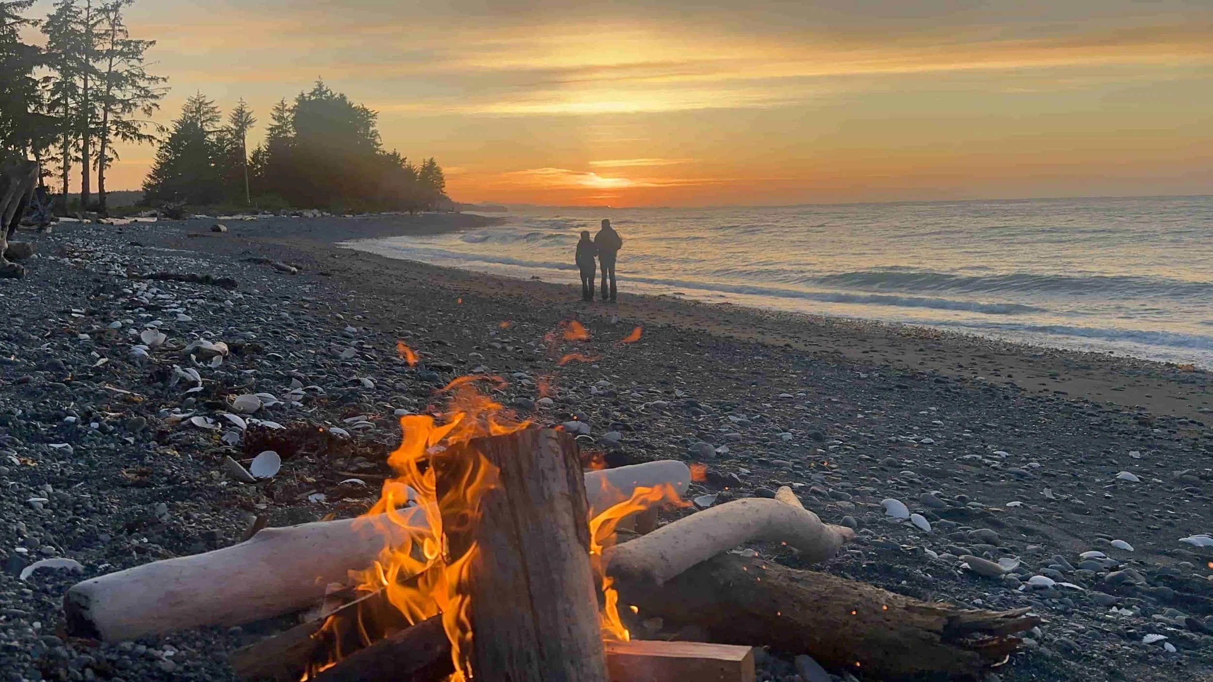 Campfire on the beach at sunset with people walking along the shore at Cluxewe Resort Campground Vancouver Island