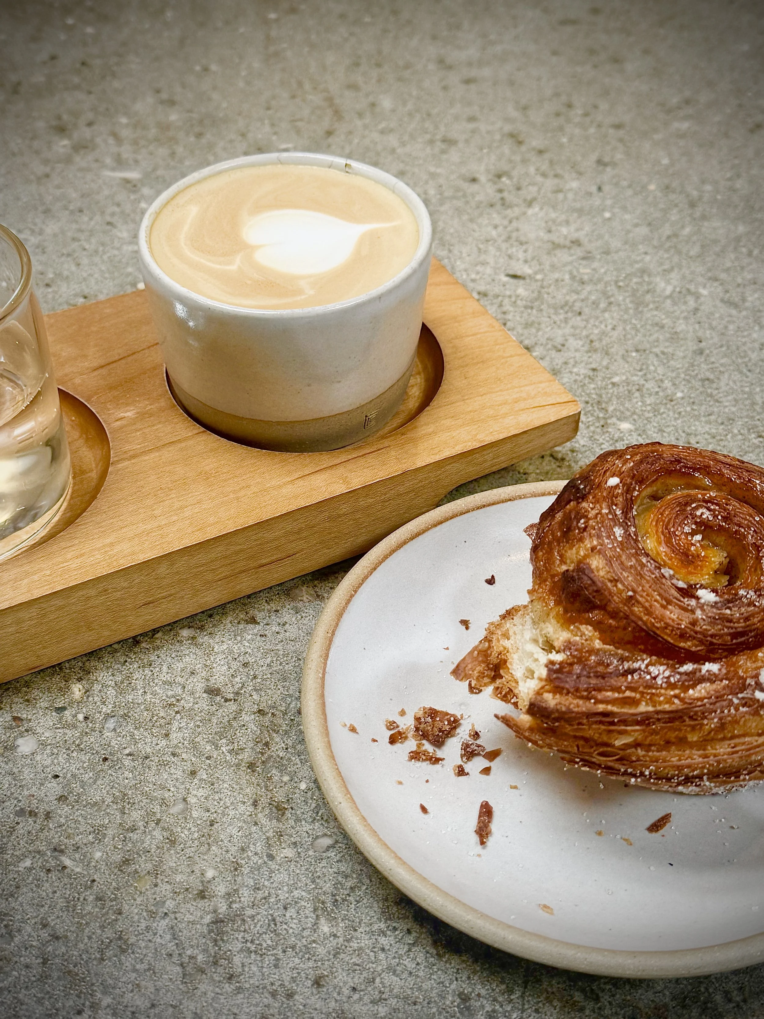 A cinnamon roll on a white plate with a few crumbs, accompanied by a cup of coffee with foam and latte art, placed on a wooden tray on a gray textured surface.
