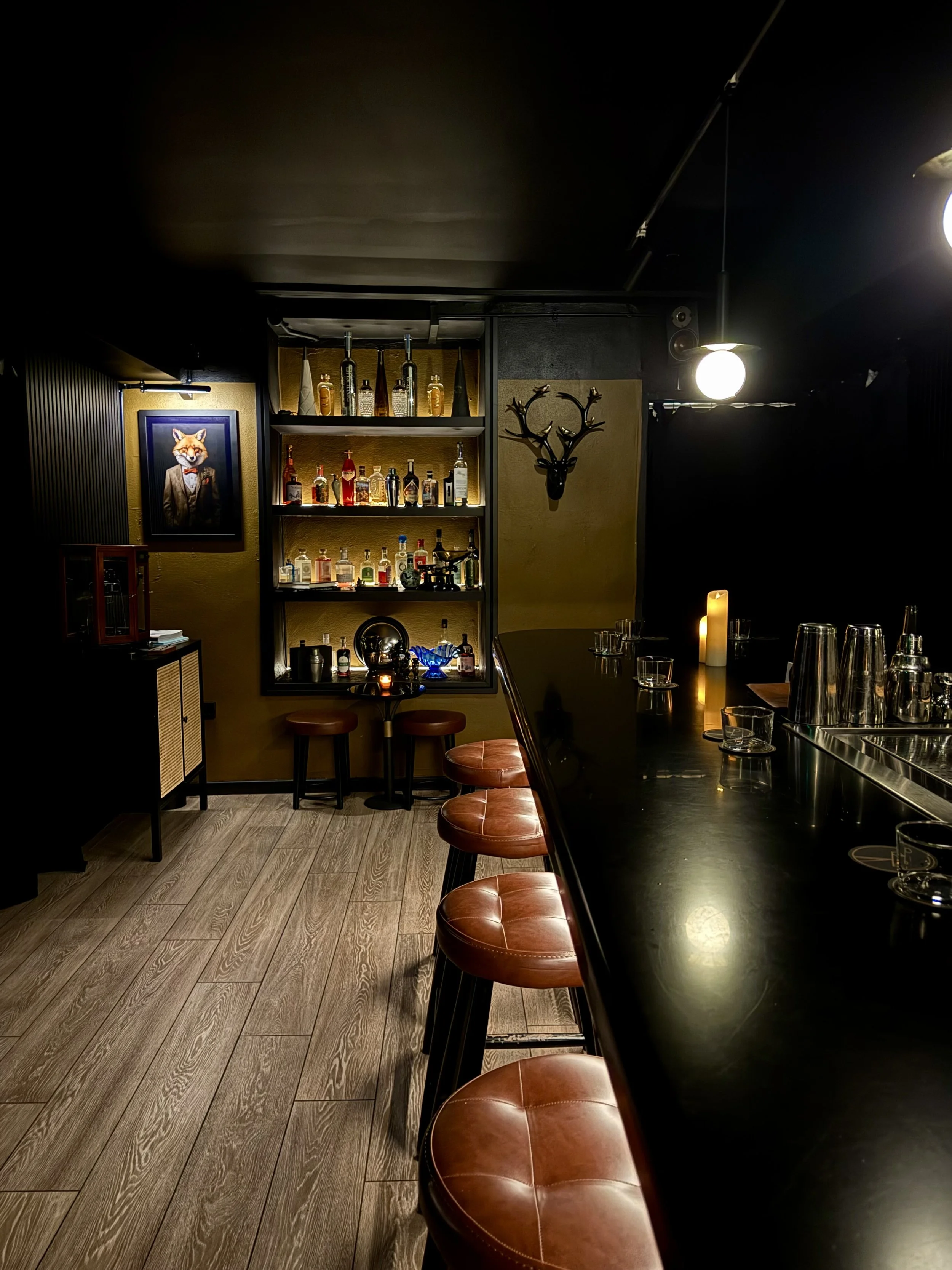 Interior of a dimly lit bar with leather stools around a black counter. Shelves with bottles of alcohol, a framed fox portrait, and a decorative deer head on the wall.