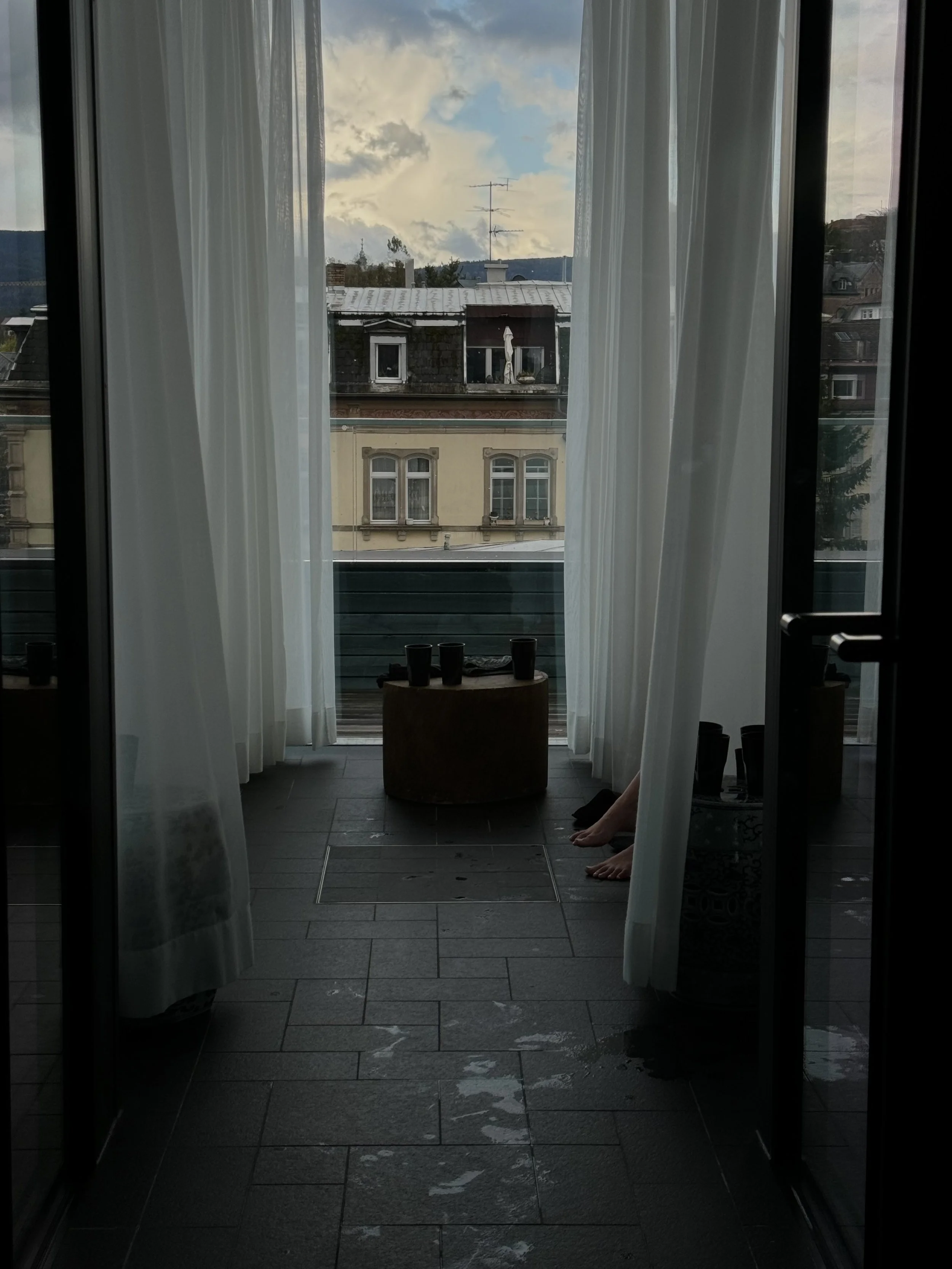 View from a balcony or patio through glass doors, with sheer white curtains, looking at rooftops and a cloudy sky with patches of blue.