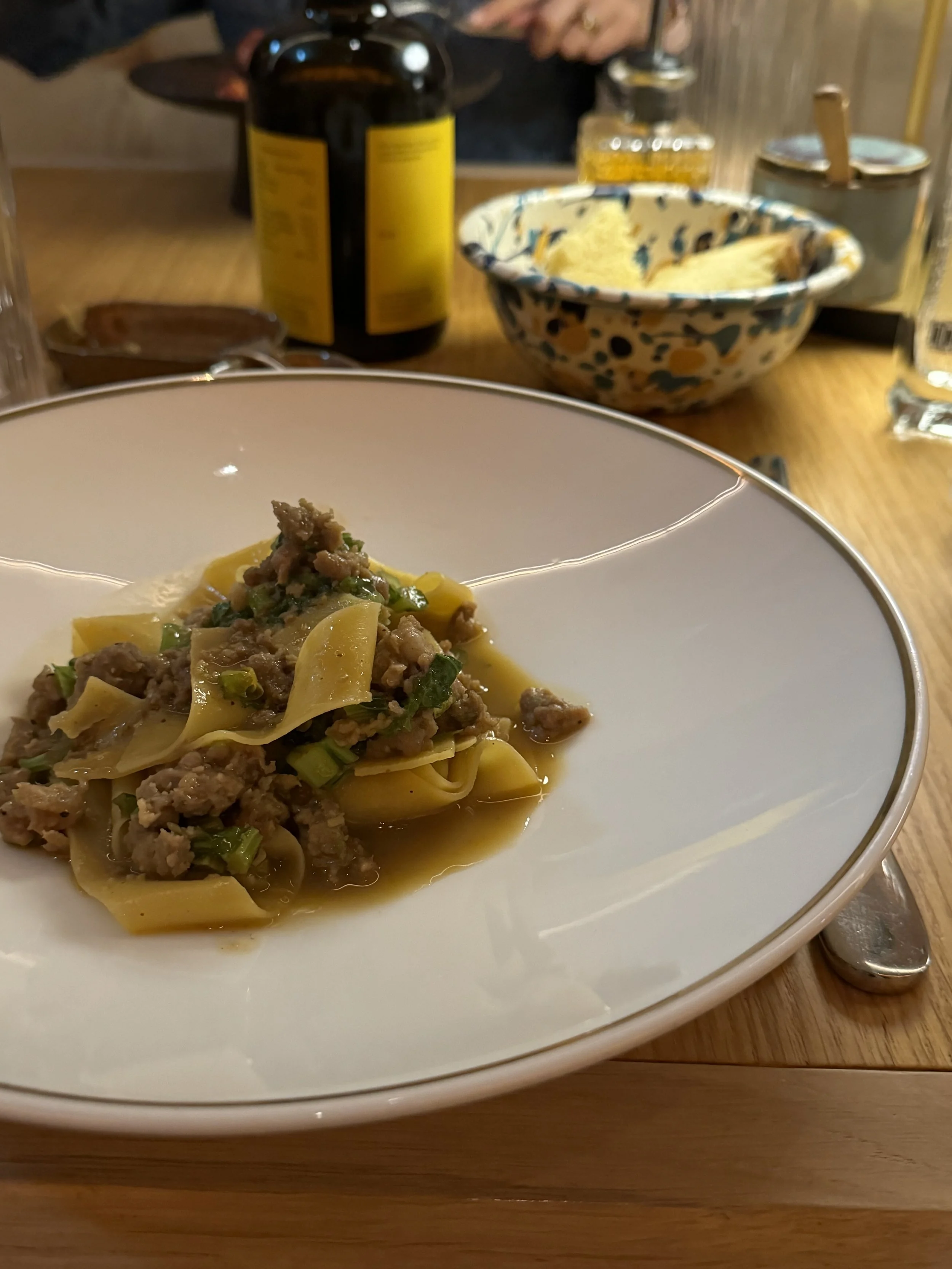 A white plate with pasta and ground meat, garnished with green onions, on a wooden table. In the background, there is a bowl of grated cheese, a bottle of drink, and various condiments.