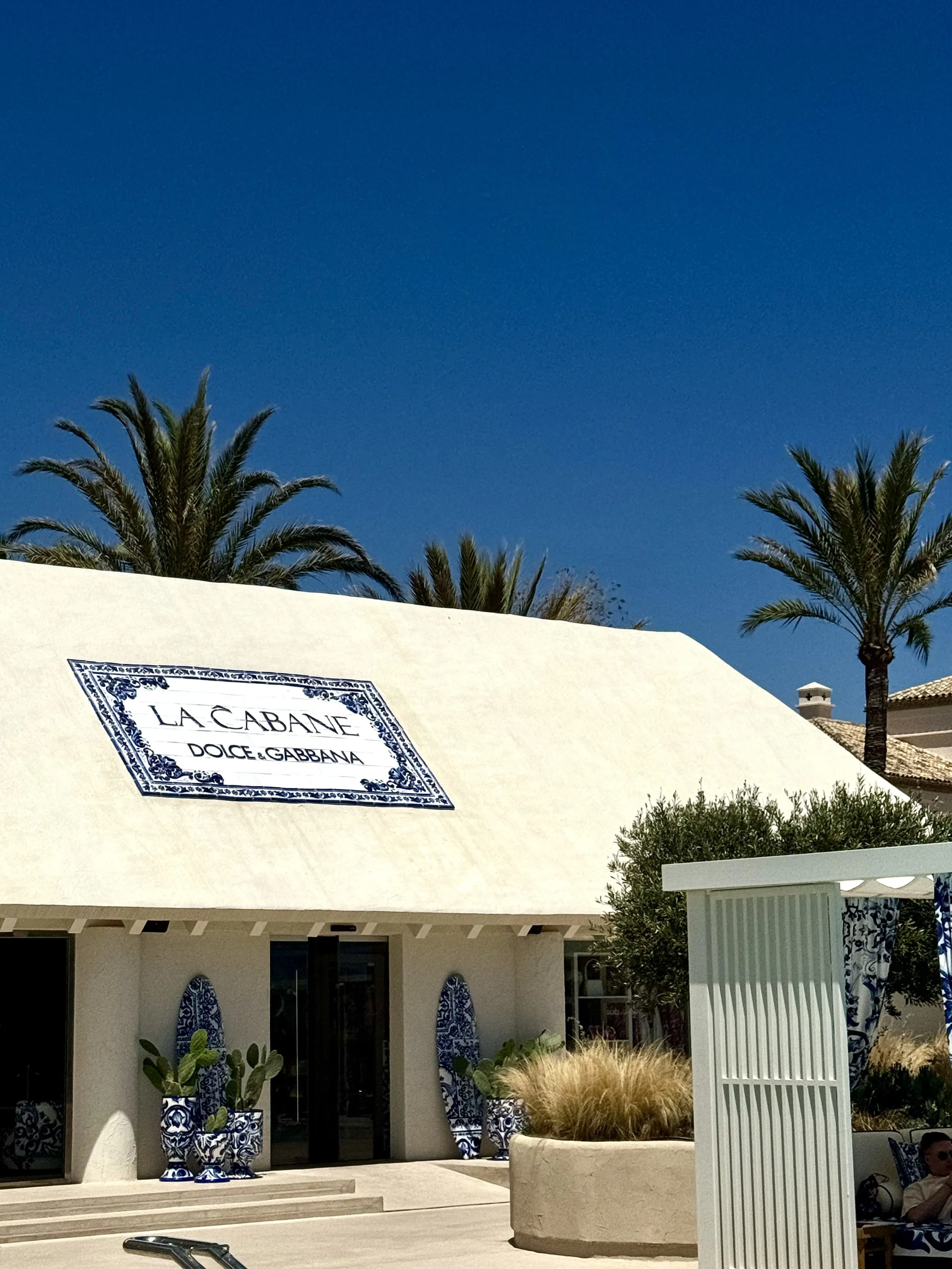 Exterior of La Cabane boutique clothing store with white awning, palm trees in the background, and blue and white decorative elements outside.