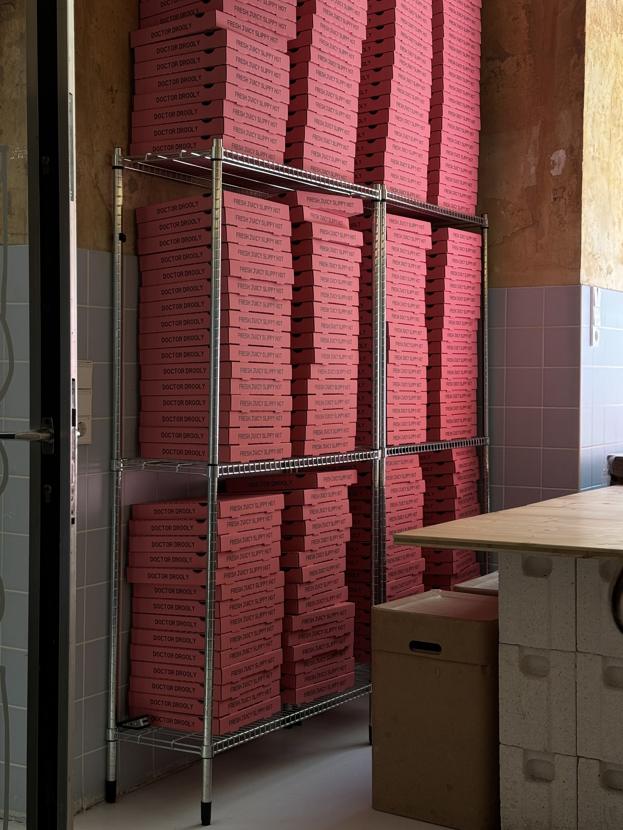 A metal shelving unit filled with stacked pink boxes labeled 'DR. DROOLY' and 'FRESH JUICY SLIPPY HOT,' situated in a room with tiled walls and a wooden counter nearby.