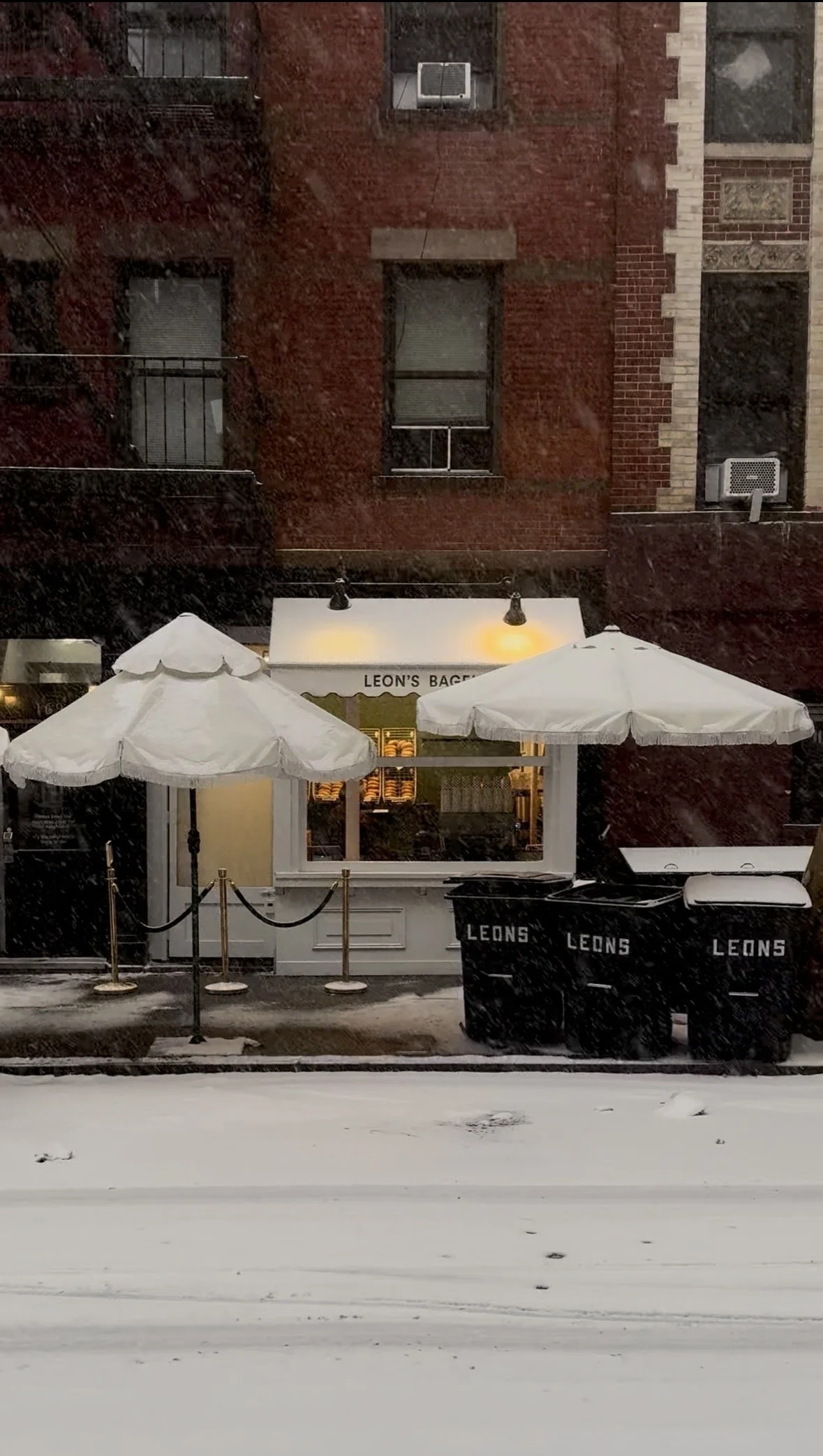 Snow-covered sidewalk and street view outside a small bakery storefront with white umbrellas, black trash bins labeled 'Leons,' and a lit window.