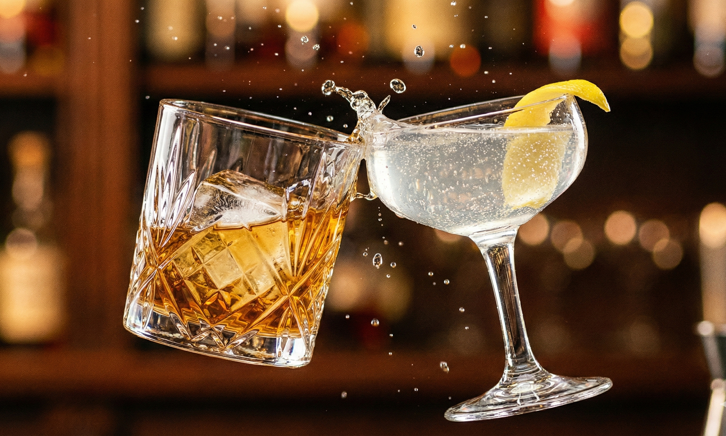 A glass of whiskey with ice clinks against a cocktail glass with a lemon wedge, causing a splash of liquid to fly into the air.