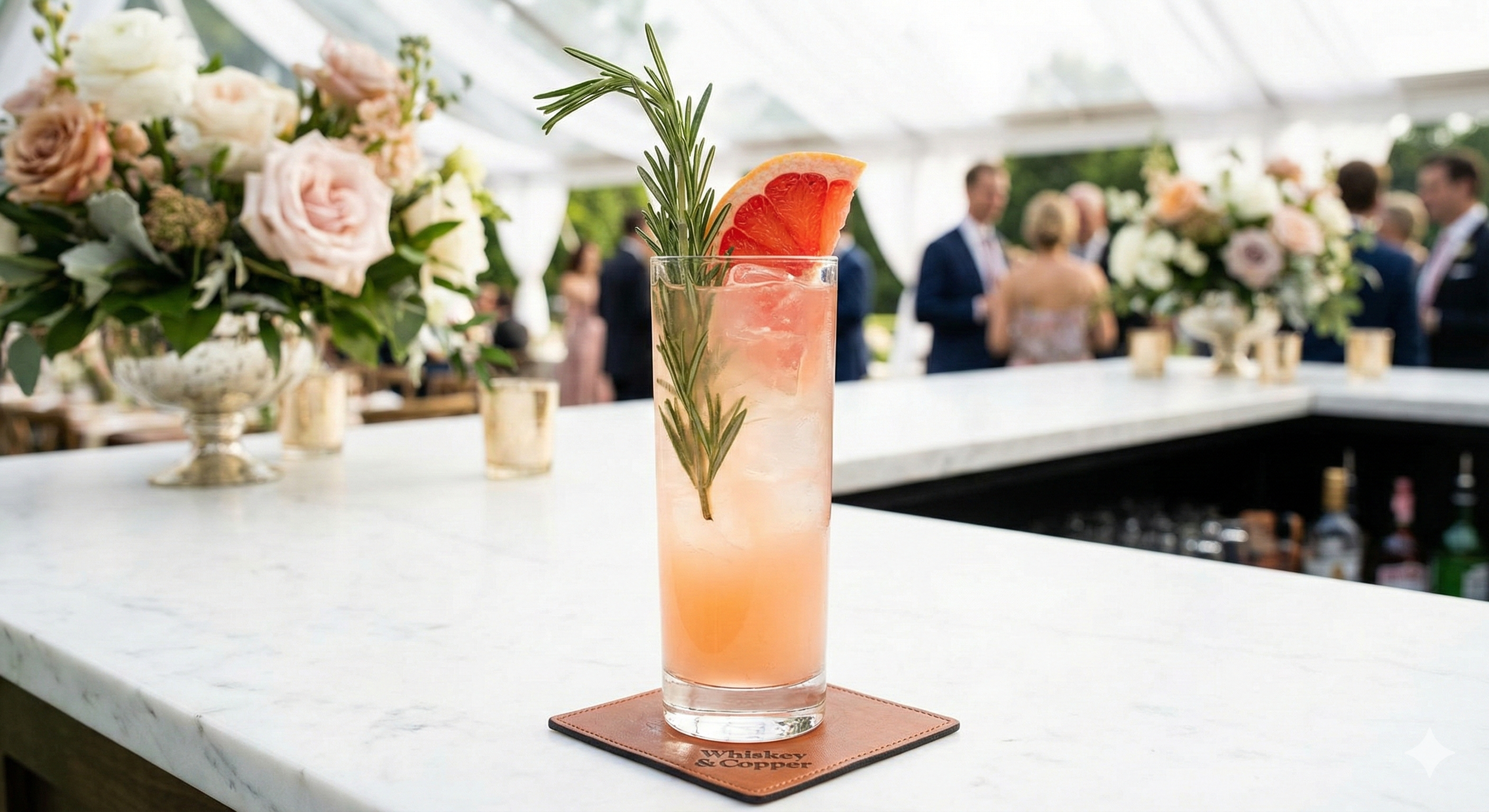 A pink cocktail garnished with a slice of grapefruit and sprigs of rosemary on a bar counter at a social event with flowers and guests in the background.