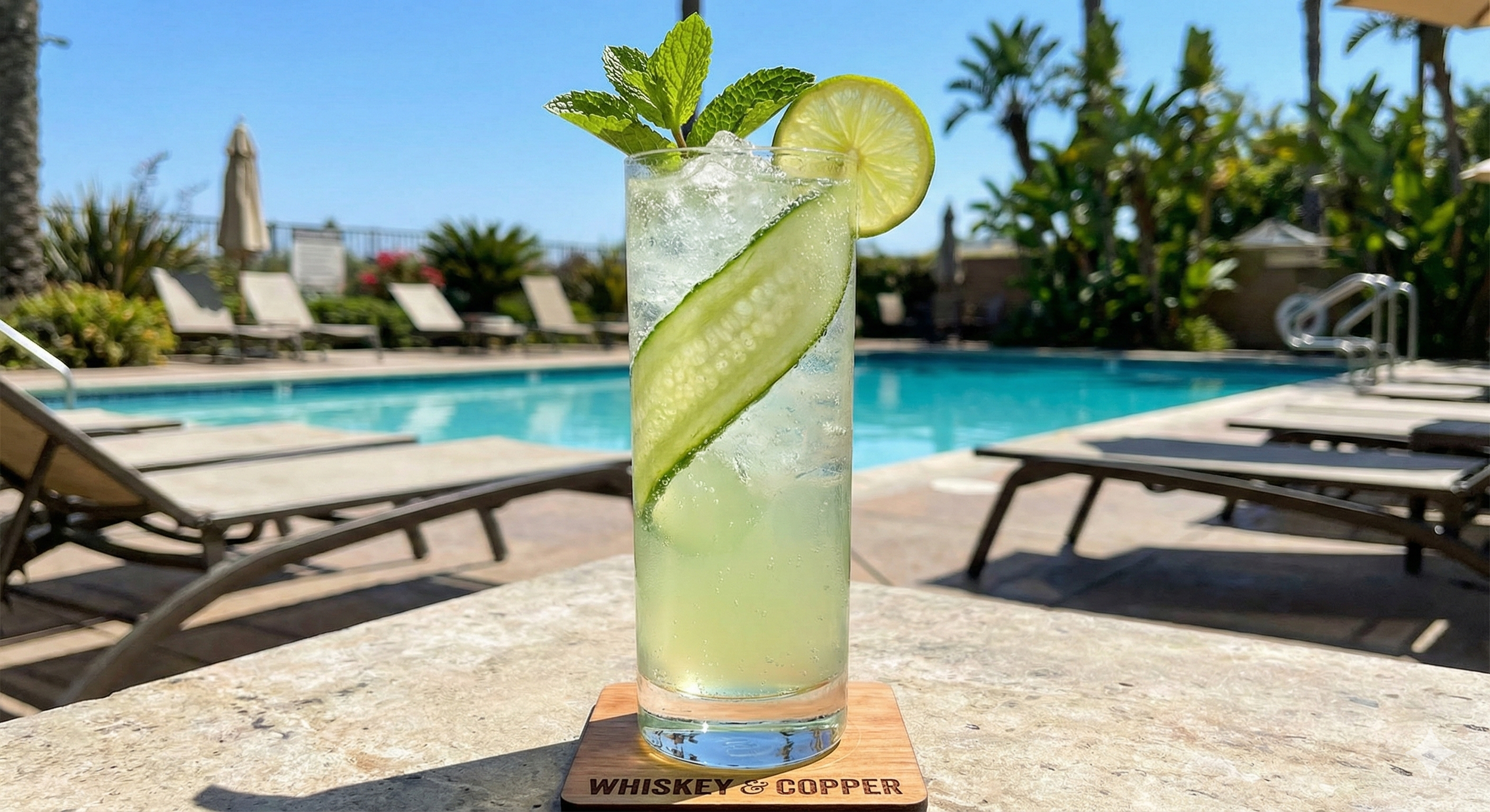 A tall glass of cucumber, lime, and mint cocktail with ice, sitting on a wooden coaster that says 'WHISKEY & COPPER' beside a pool outdoors on a sunny day.