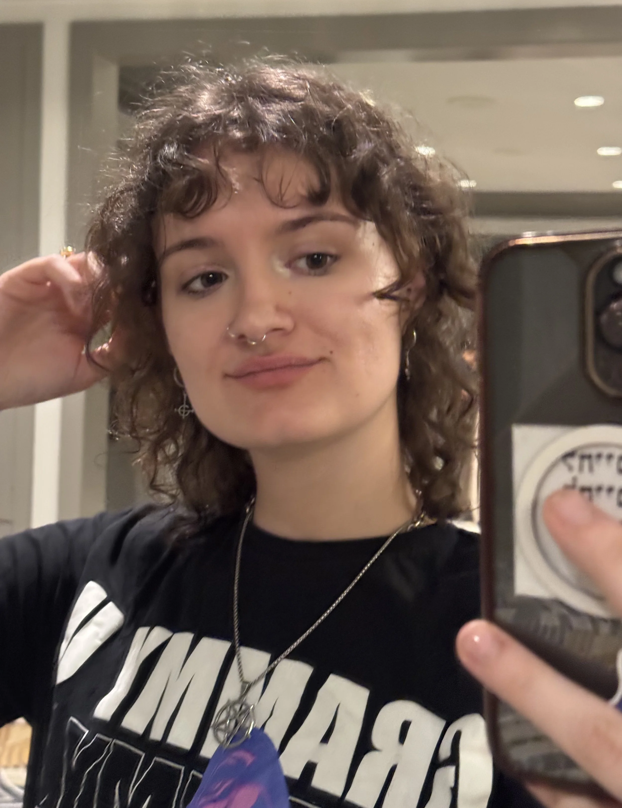 A young woman with curly hair taking a mirror selfie in a room with overhead lighting. She is wearing a black T-shirt with white text, a necklace, and earrings. She has makeup, a nose ring, and is looking at her phone's camera with a slight smile.