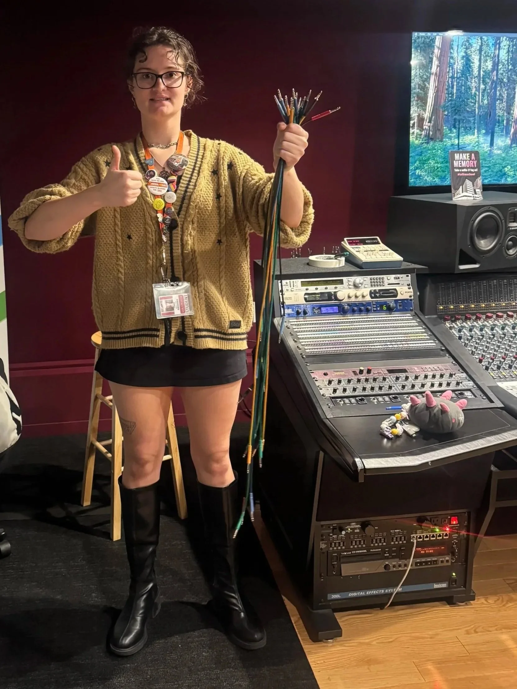 A woman holding a bow and arrows, giving a thumbs-up in a music studio with audio equipment, speakers, and a monitor displaying a forest scene.