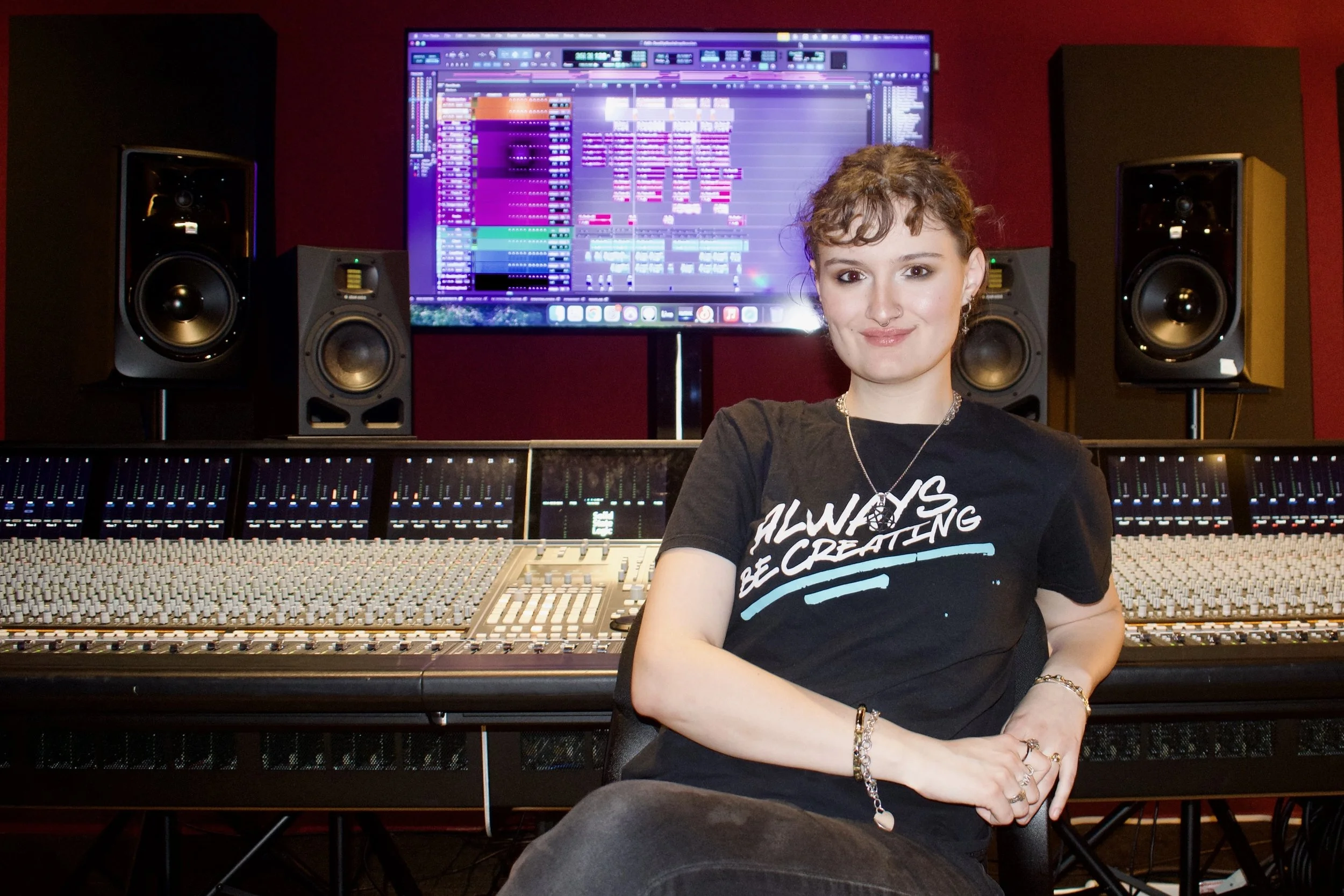 Young woman with short curly hair sitting in a music studio in front of mixing console and speakers, wearing a black T-shirt that says 'Always Be Creating'.