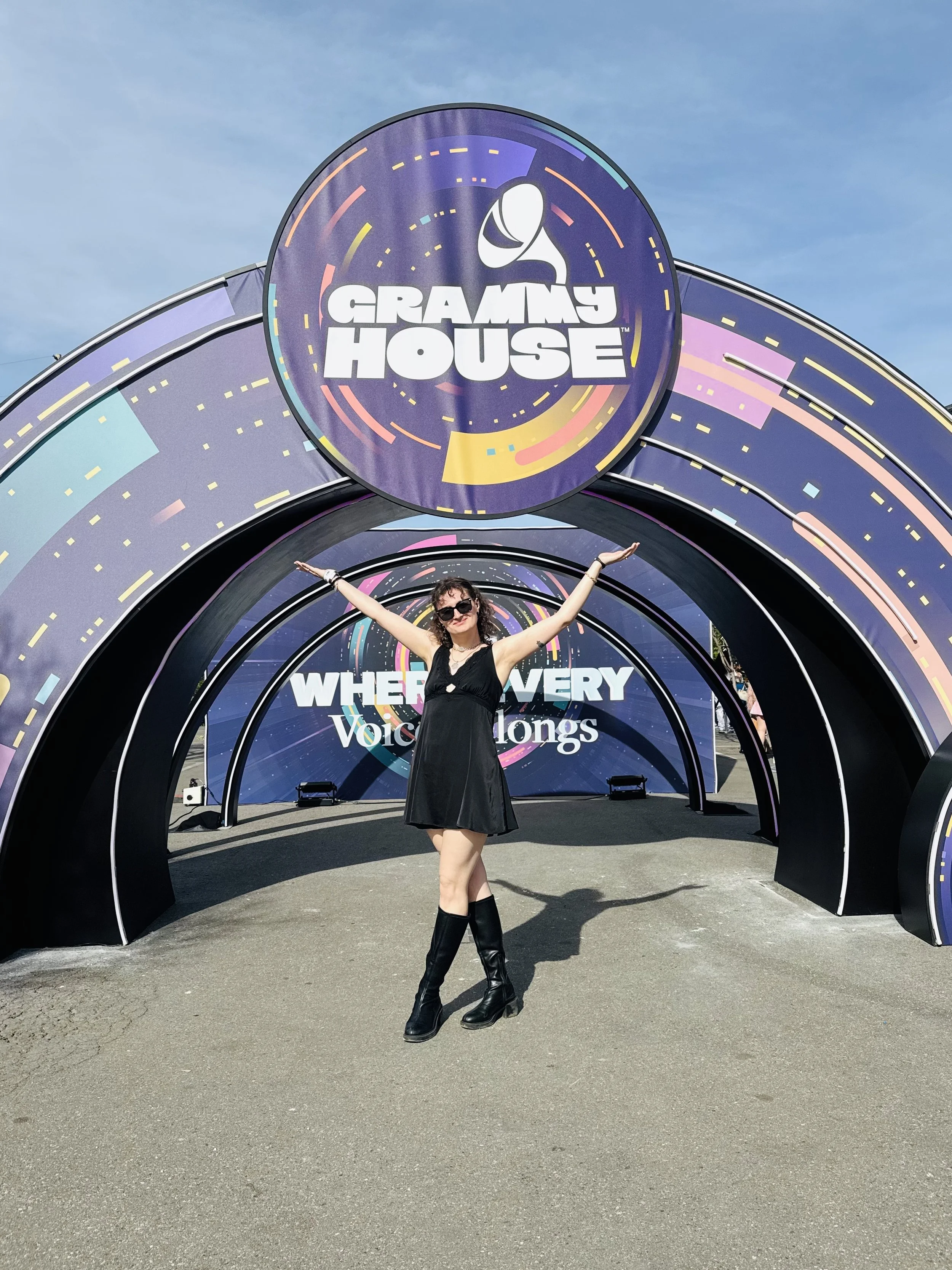A woman is standing with arms outstretched in front of a colorful stage display with the logo of Grammy House at the top and the words 'Where Every Voice Matters' behind her.