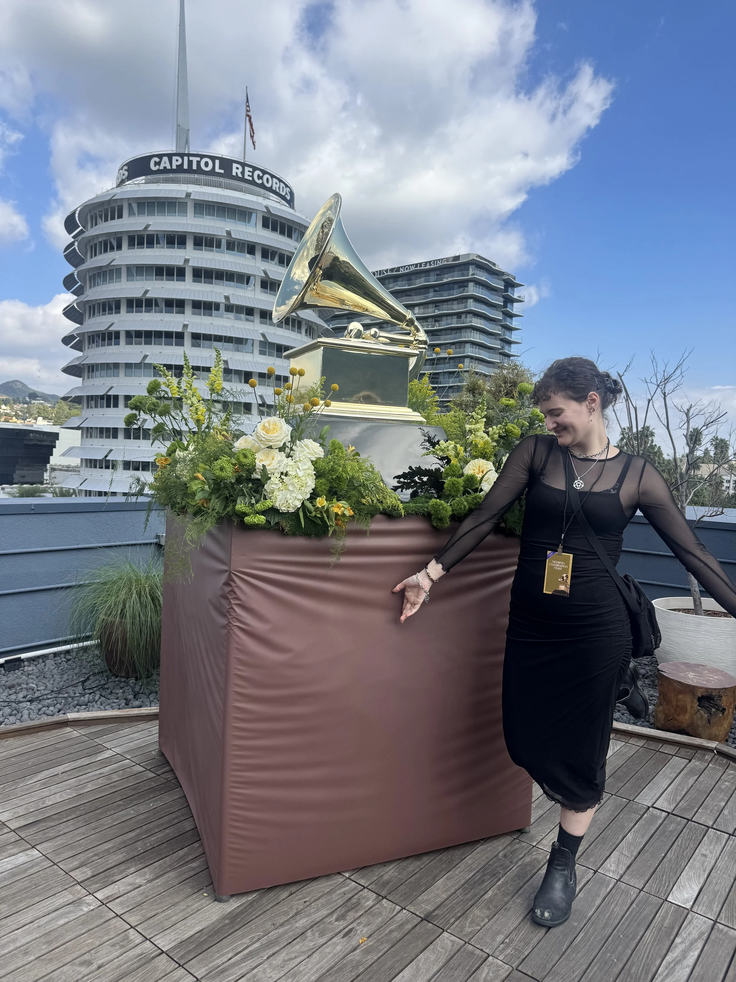 A woman in a black dress and boots smiling and pointing at a large vintage gramophone with a flower arrangement on a rooftop terrace, with the Capitol Records building in the background and a partly cloudy sky.