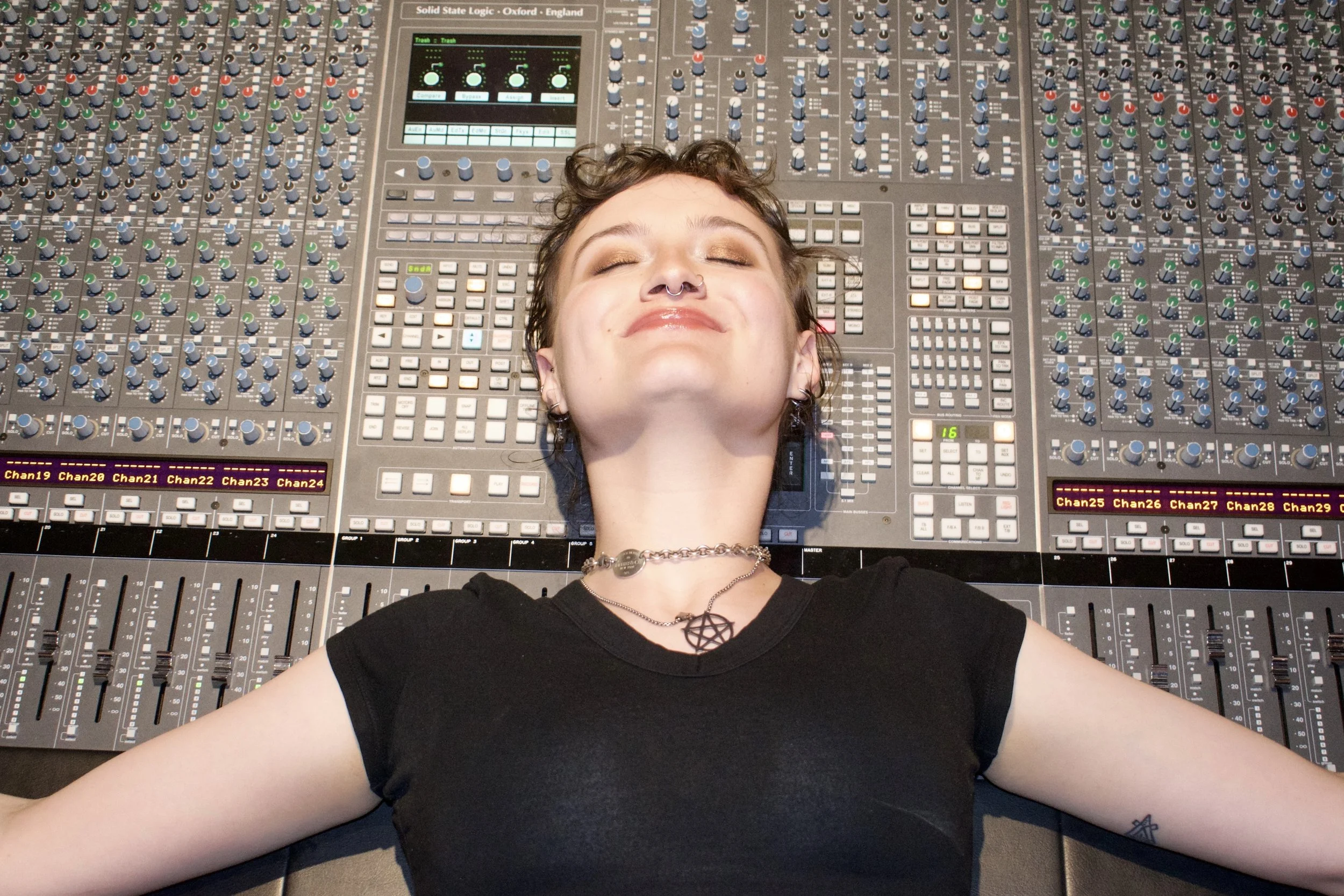 A young woman with short hair, wearing a black t-shirt, lying back with her arms spread out, resting her head on a large audio mixing console, smiling with her eyes closed.