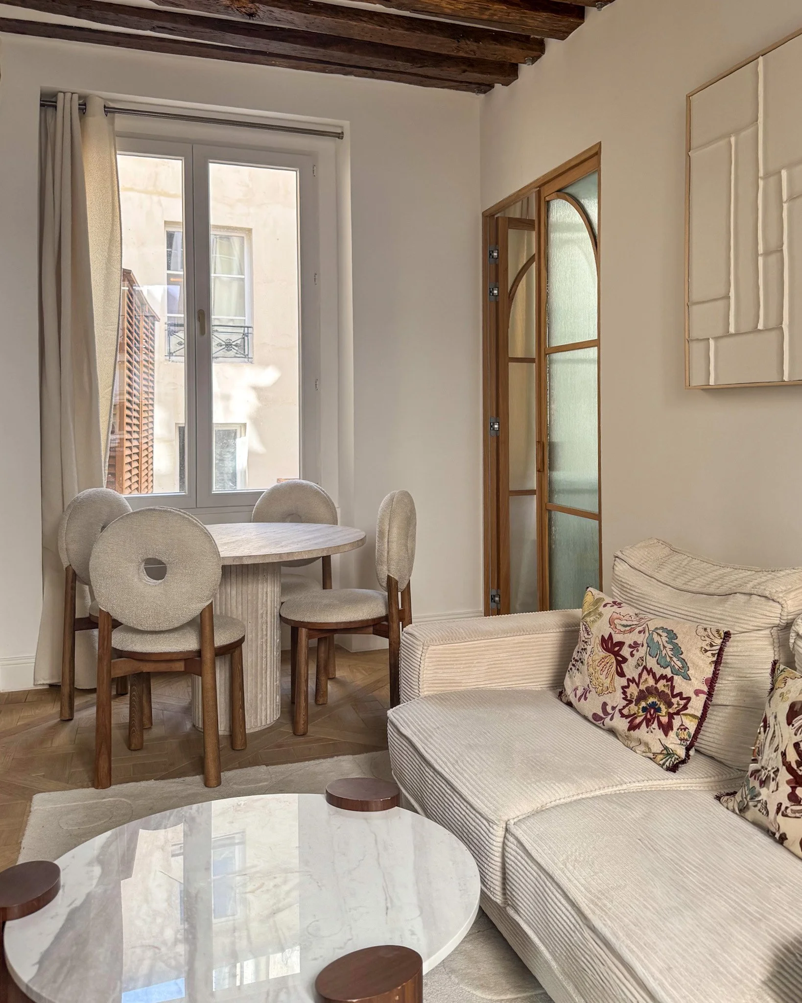 Living room with a large window, beige couch with floral pillows, marble coffee table, wooden dining table with chairs, and wood-beamed ceiling.