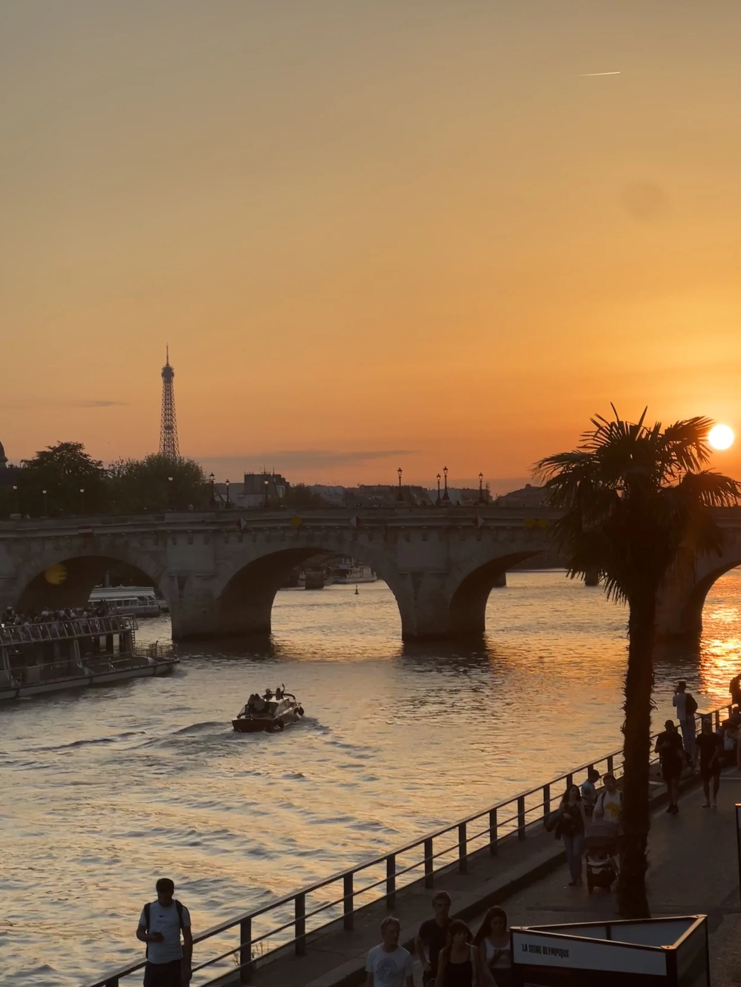 Sunset over the Seine River in Paris, with the Eiffel Tower visible in the distance, people walking along the riverbank, and boats on the water.