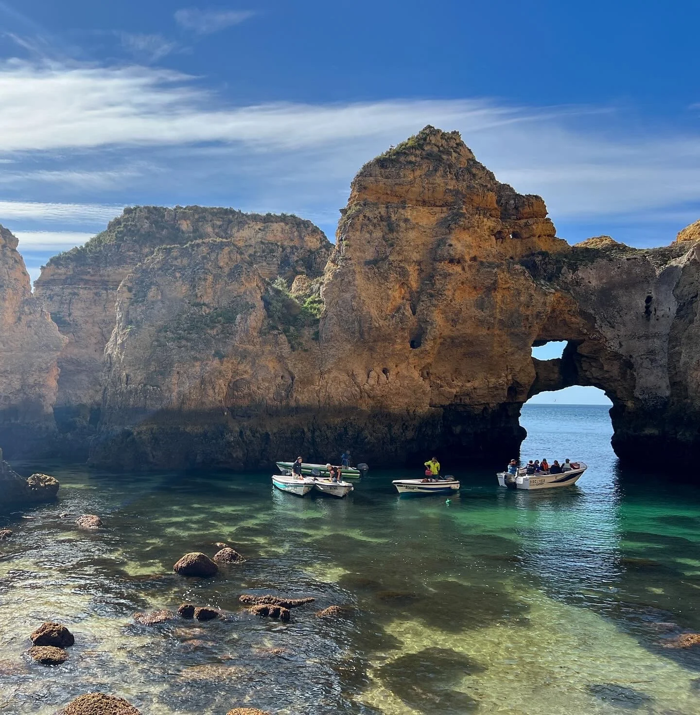 Cliffs with a natural arch formation over a calm body of water, with several boats carrying people near the rocks.