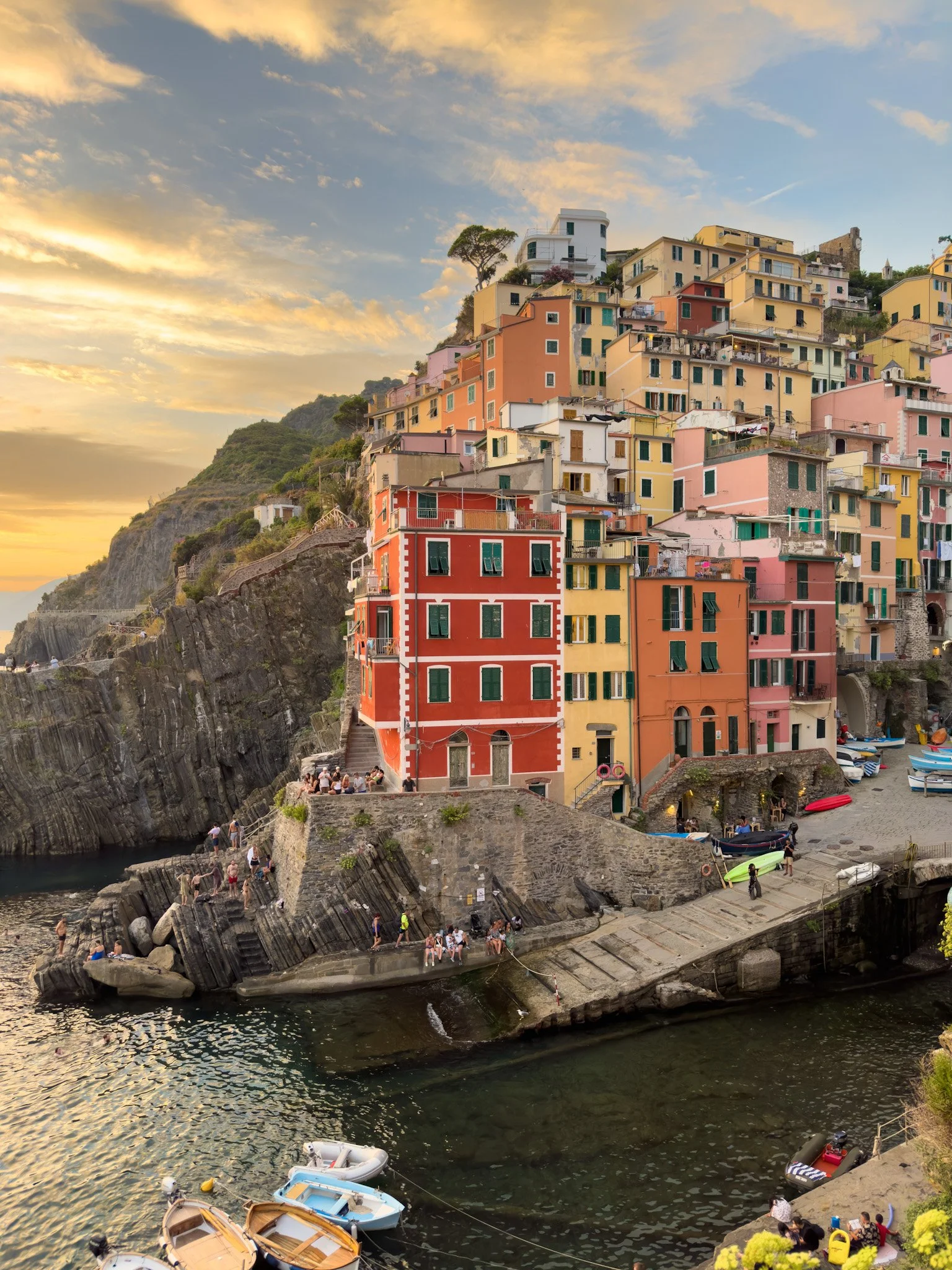 Colorful houses built on a hillside above a harbor at sunset, with boats docked in the water and people walking on the rocks and stairs.