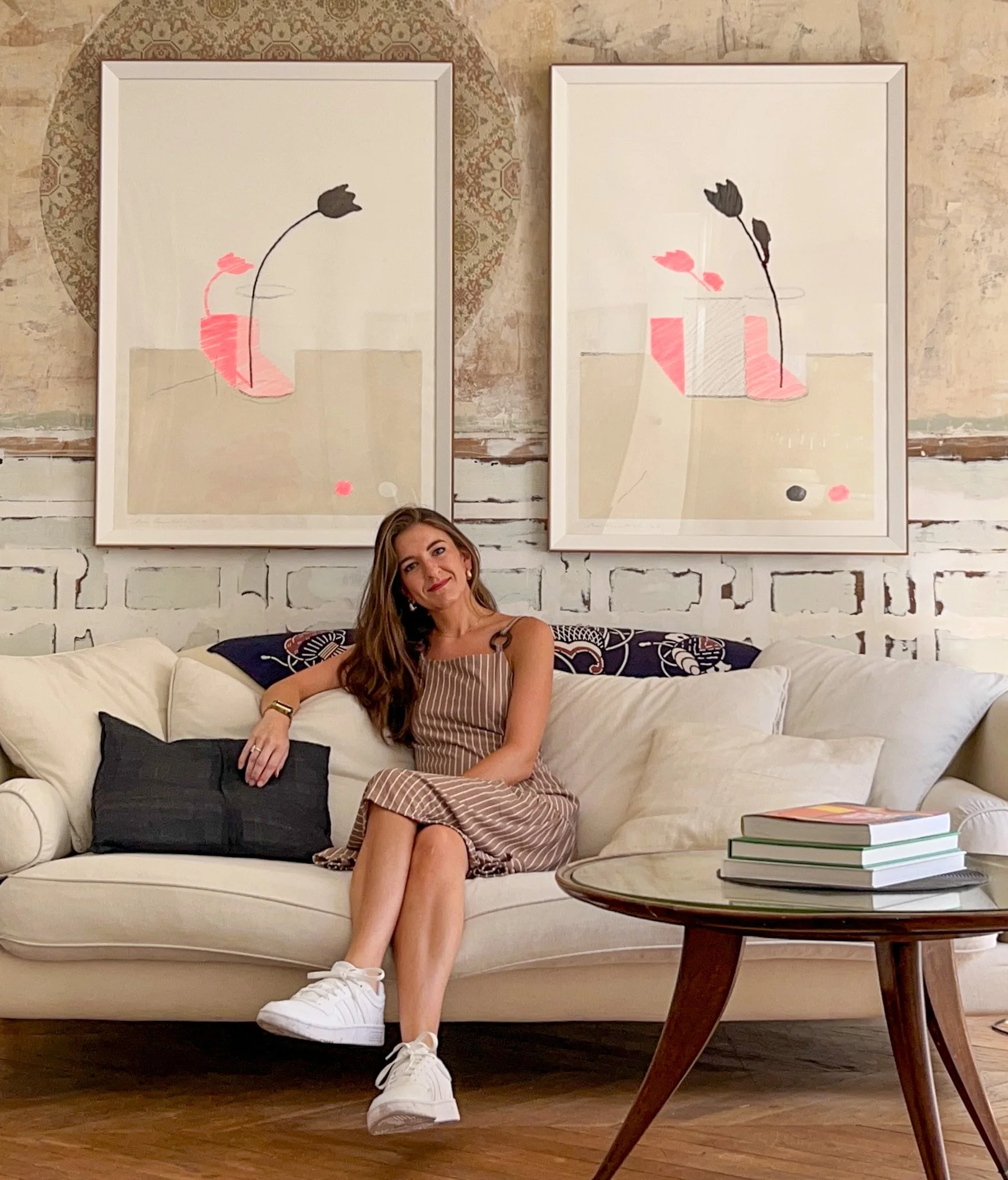 A woman sitting on a white couch in a cozy, art-filled living room, looking at the camera with a relaxed smile.