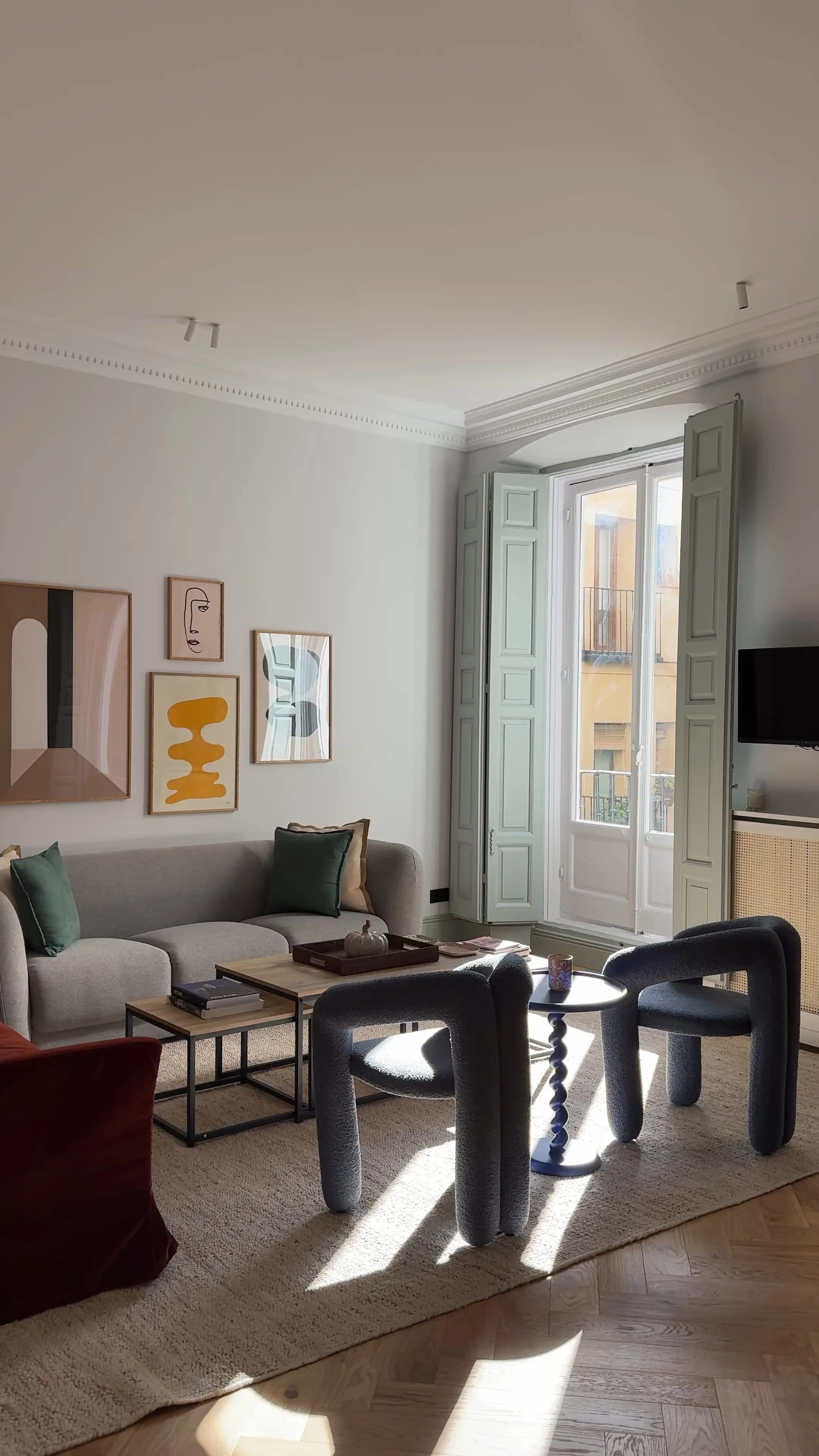 Living room with a beige couch, colorful framed artwork on the wall, a window with open shutters allowing sunlight, a small side table with a mug, and a television mounted on the wall.