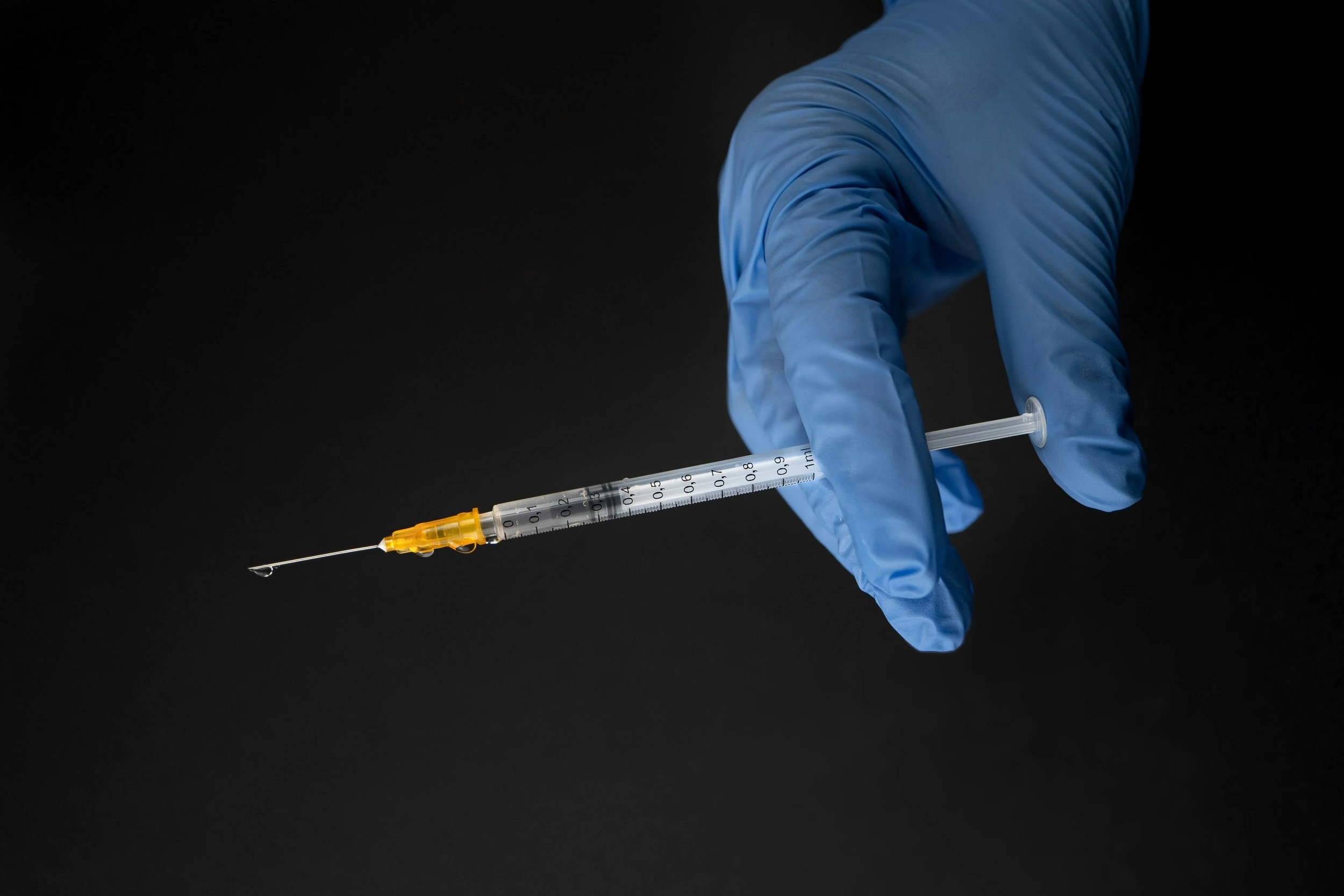 Una mano con guanto blu tiene una siringa con ago sottile contro uno sfondo nero.