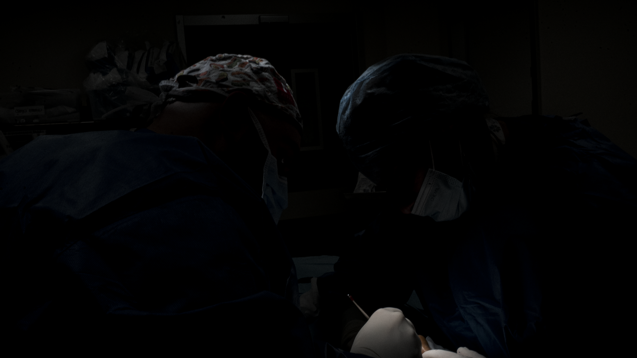 L'immagine ritrae due persone, probabilmente medici o infermieri, durante un intervento chirurgico in un ambiente sterile, con luci concentrate sull'area di lavoro.