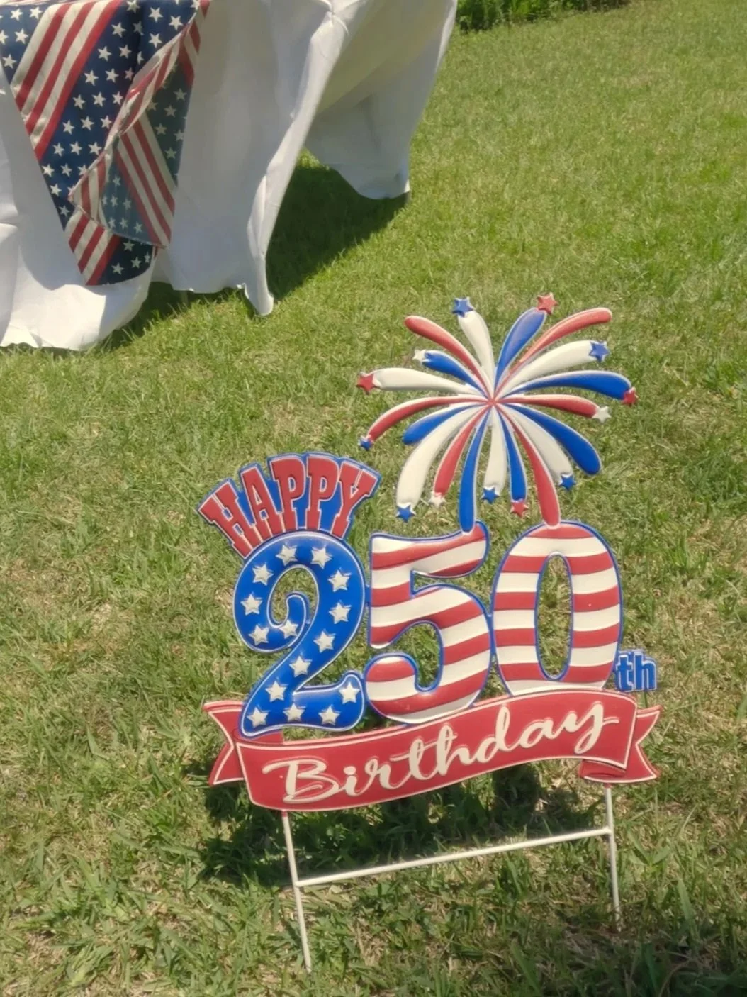 Red, white, and blue birthday sign with the number 250, a firework decoration, and a sign reading 'Happy 250th Birthday' on green grass with part of a table draped with American flag-themed cloth in the background.
