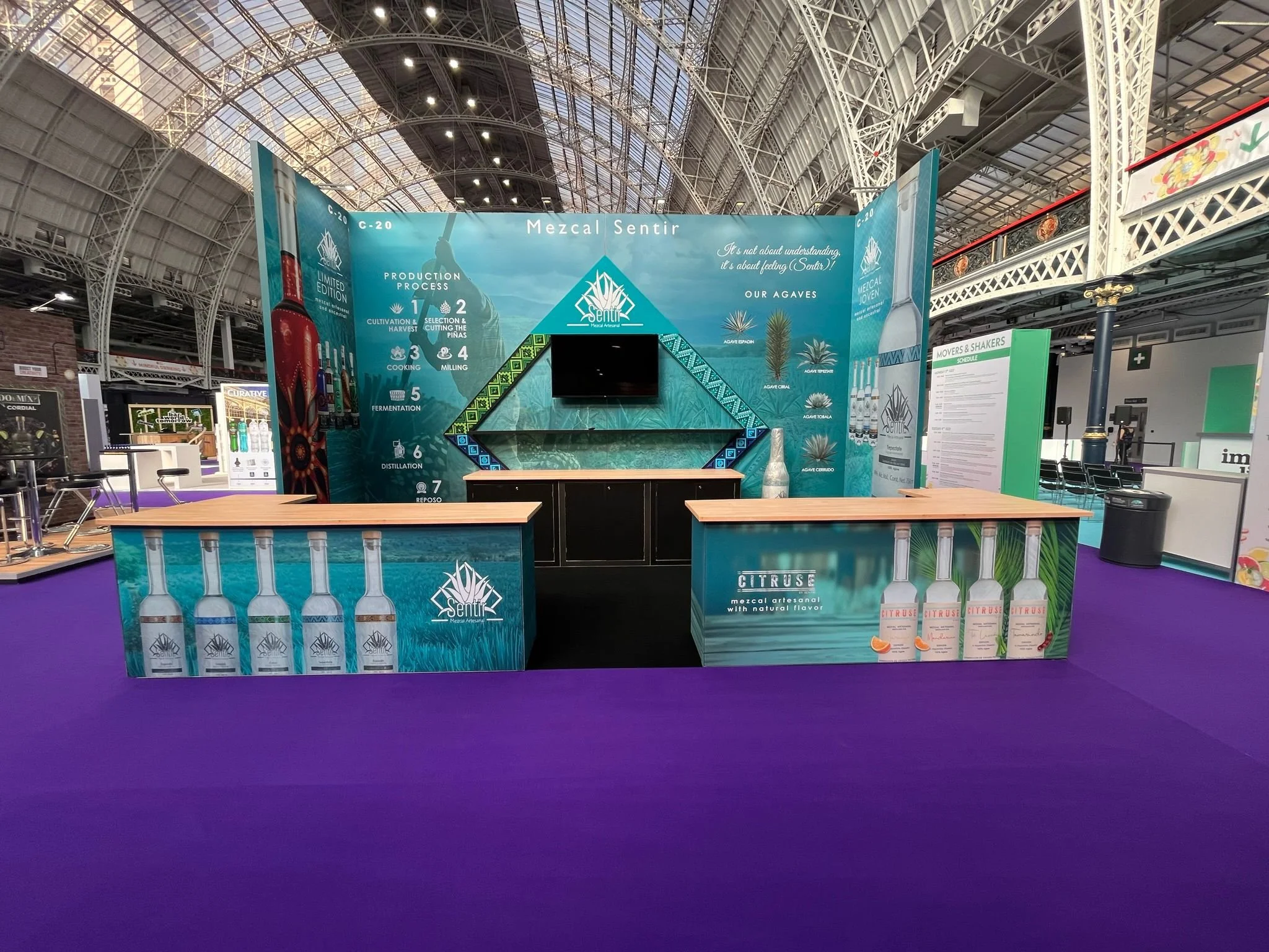 Exhibition booth showcasing mezcal products with colorful branding, a backdrop explaining the production process, and displays of mezcal bottles, set on a purple carpet.