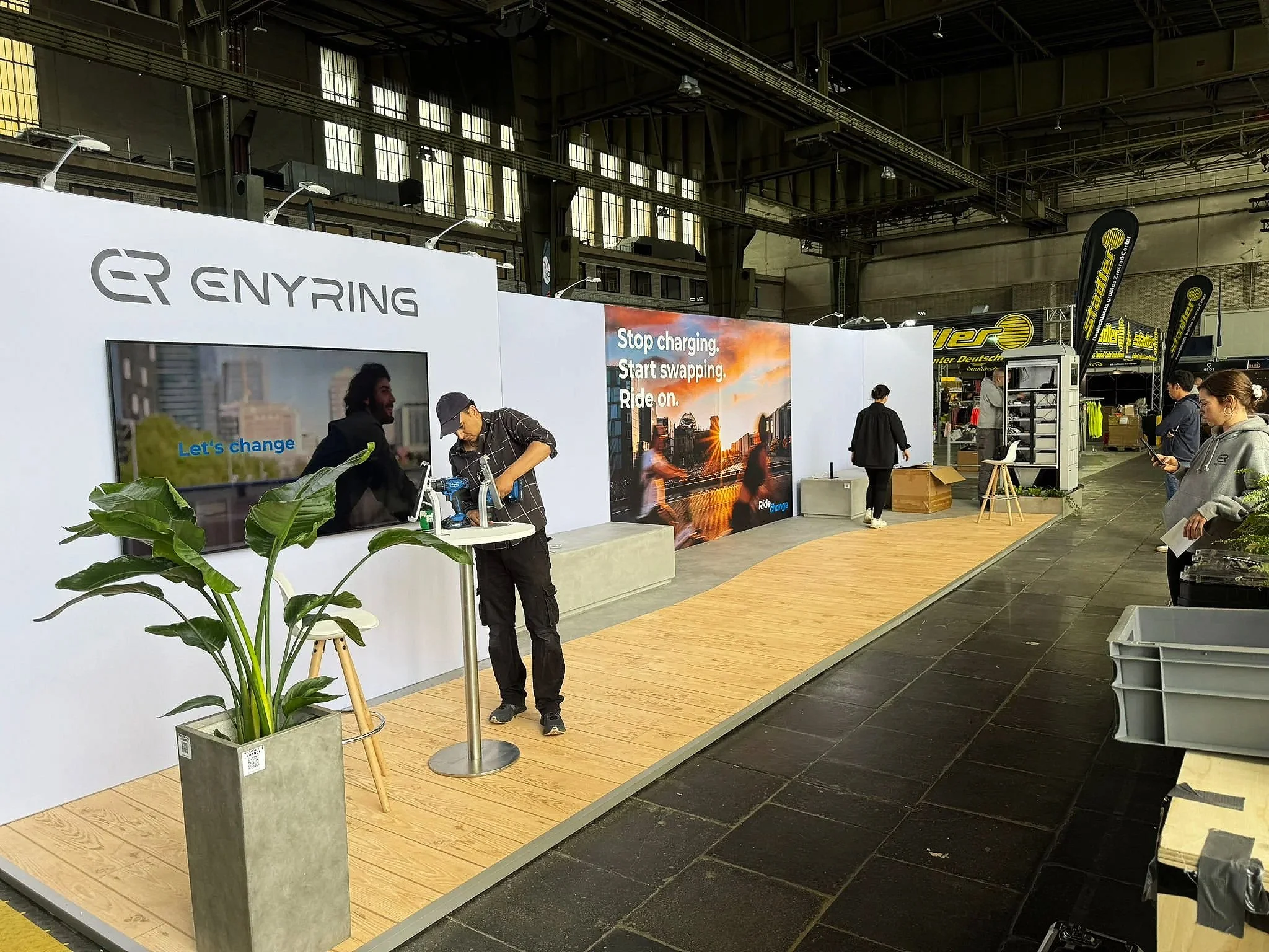 Exhibition booth for ENYING with a large screen, promotional posters, and banners, set up inside an industrial-style venue with several people around.