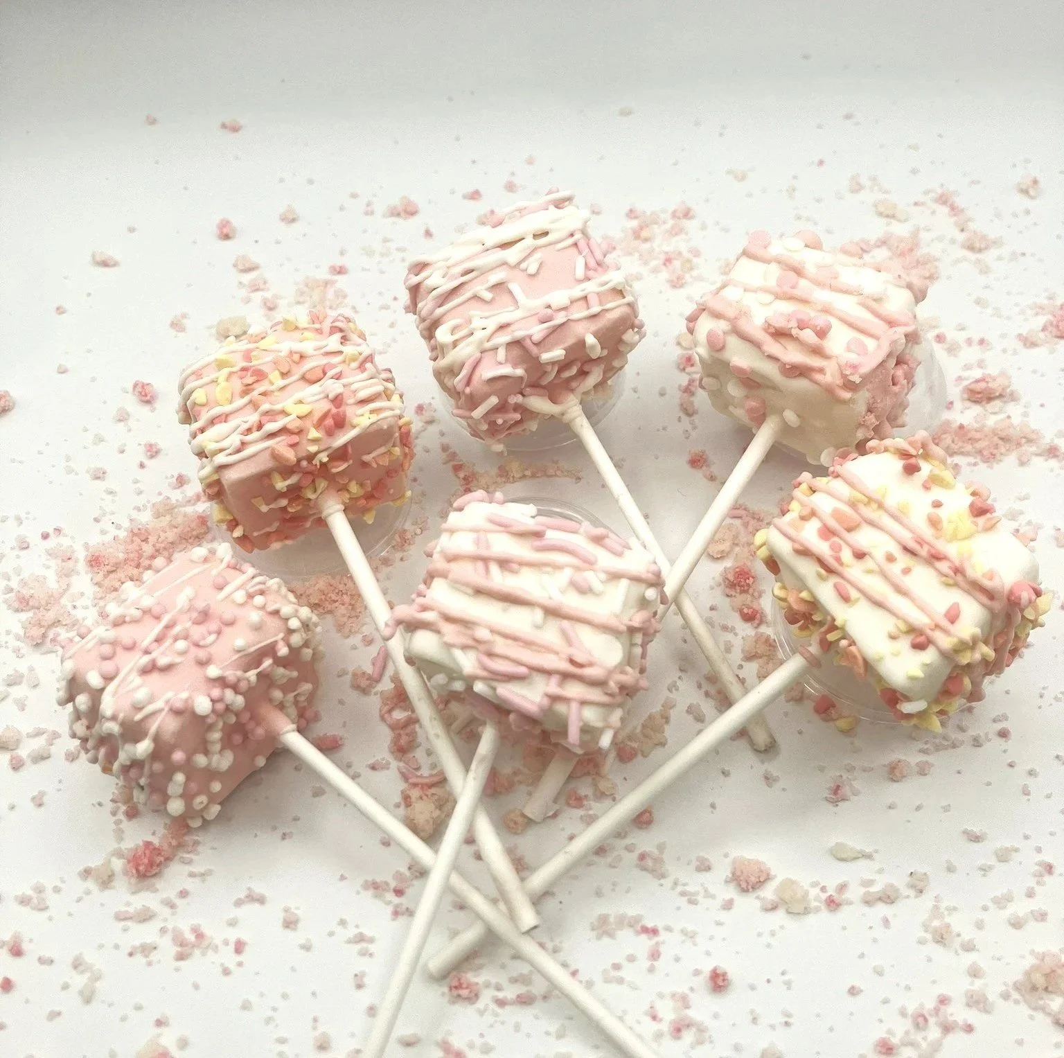 Six pink and white chocolate-covered marshmallow pops topped with pastel sprinkles, arranged on a white surface with scattered sprinkles.