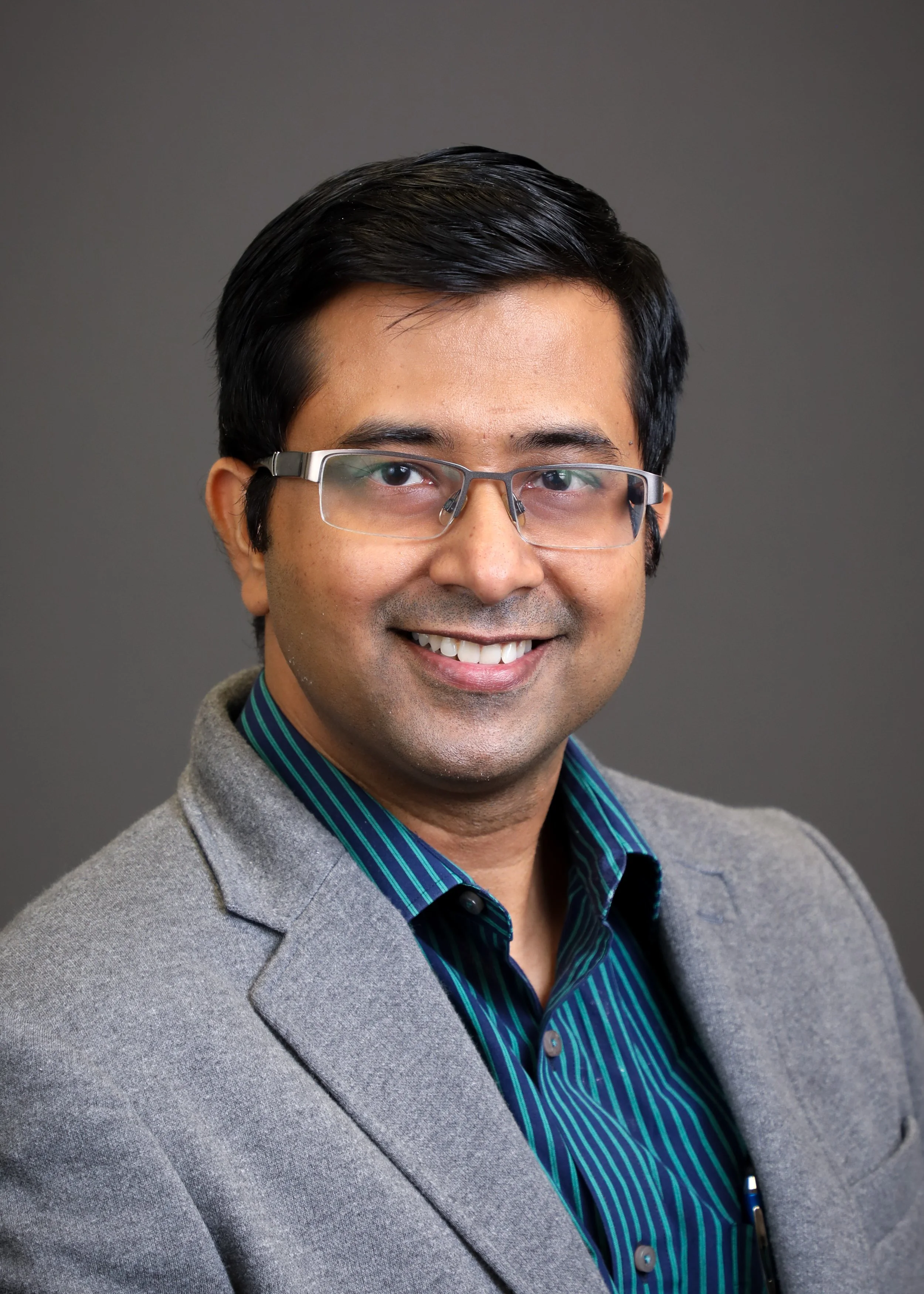 Portrait of a smiling man wearing glasses, a grey blazer, and a blue-striped dress shirt against a grey background.