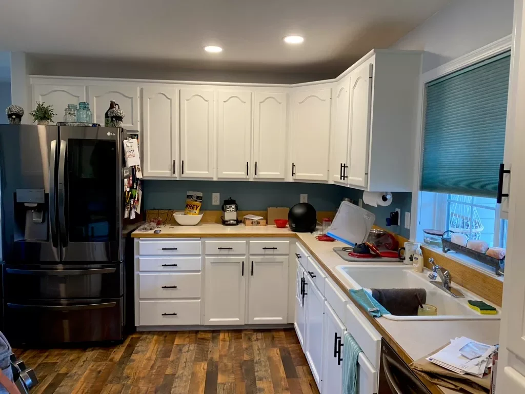 Kitchen with white cabinets, stainless steel refrigerator, and wooden countertop. The sink area contains a towel, sponge, soap dish, and papers. The window above has a closed green blind.